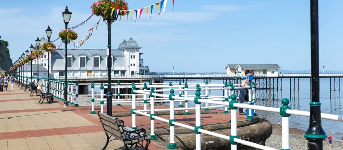 Penarth promenade with benches, black lampposts decorated with flower baskets and colorful bunting, a white building on stilts over the water, a woman leaning on the railing, and a few other people enjoying the view.