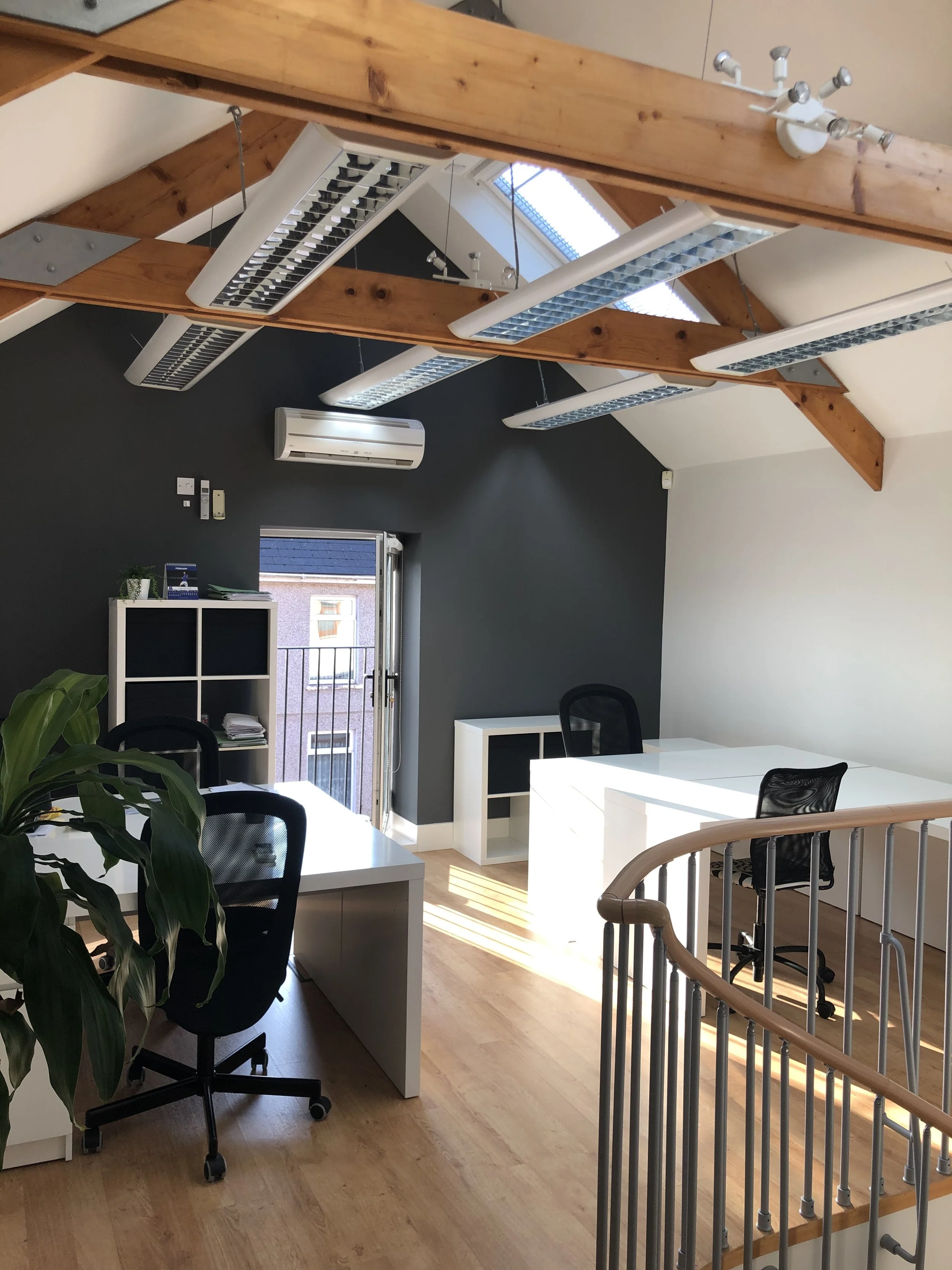 An office space with several white desks and rolling chairs, large windows allowing natural light, ceiling-mounted air conditioning, and fluorescent lights suspended from the wooden beams on the ceiling.