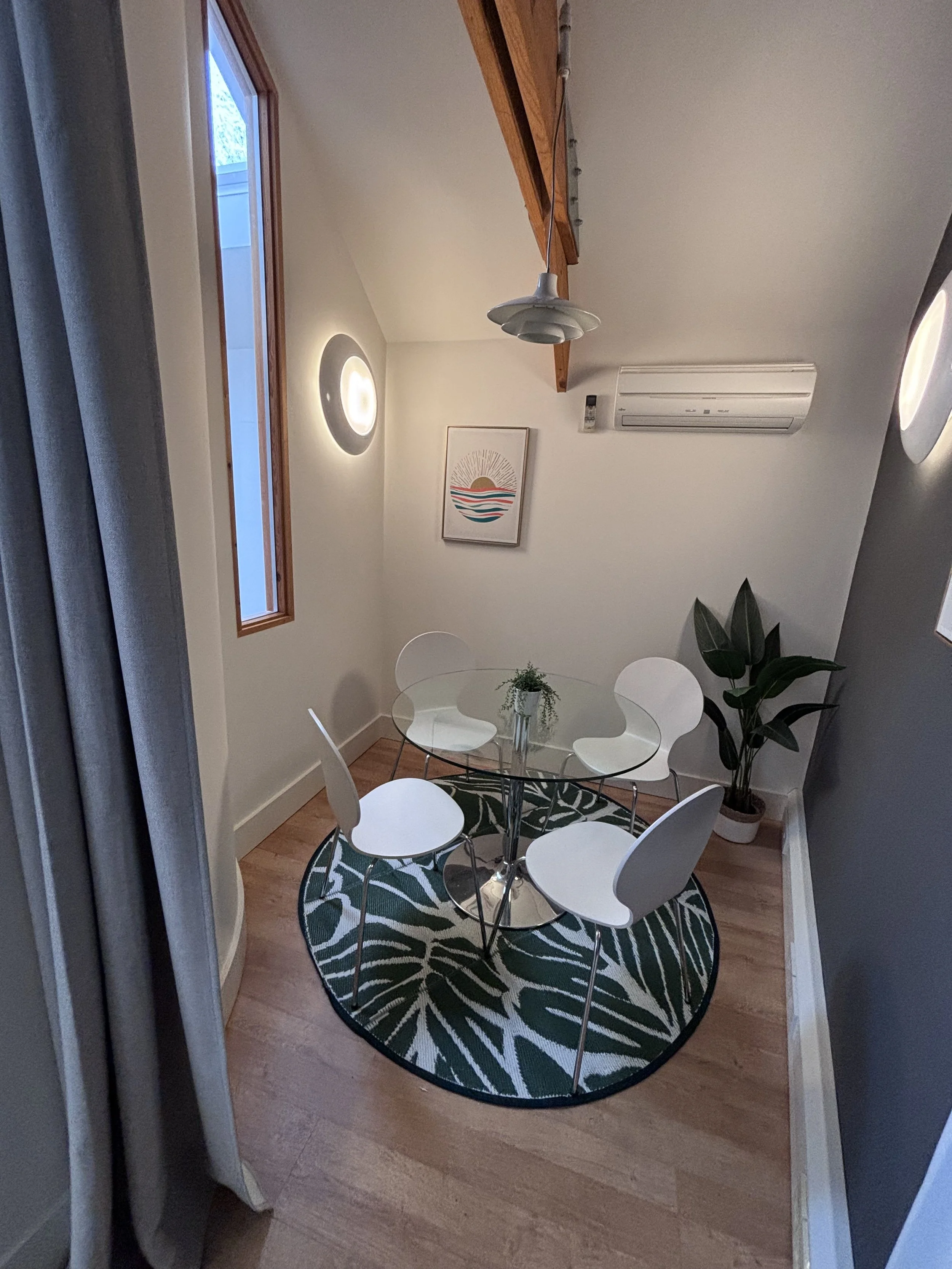 A small meeting area with a round glass table, five white chairs, a dark green and white patterned oval rug, a potted plant in the corner, a wall art depicting a sunset over water, a window, a ceiling light, and an air conditioning unit.