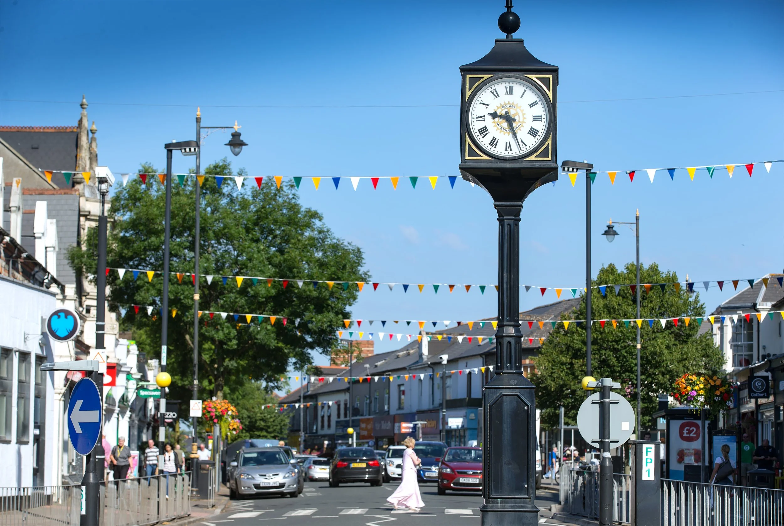 Penarth town centre, street scene with a black clock on a post displaying the time as 2:55, colorful pennant banners overhead, cars parked along the street, people walking, and buildings in the background against a clear blue sky.