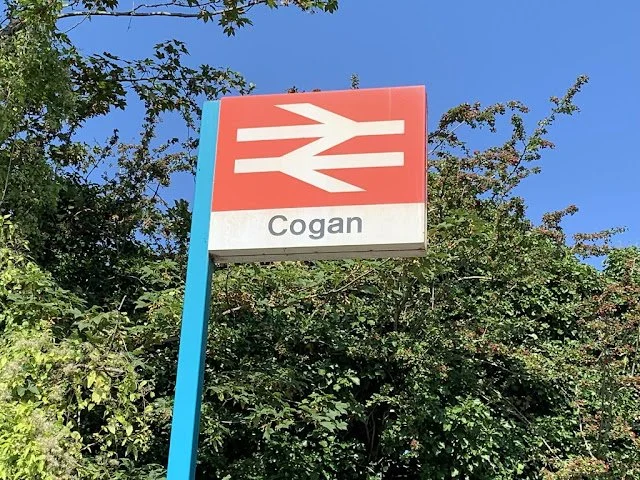 Sign for the Cogan train station with the British rail logo in red and white, blue support pole, surrounded by green trees against a bright blue sky.