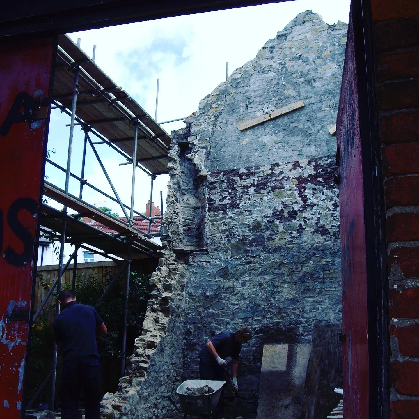 Two workers are restoring an old stone wall inside a building, with scaffolding set up nearby and a man standing in the foreground.