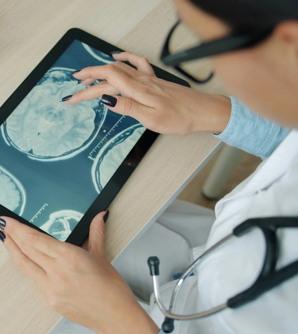 A healthcare professional in a white coat with a stethoscope around their neck is examining brain scan images on a tablet.