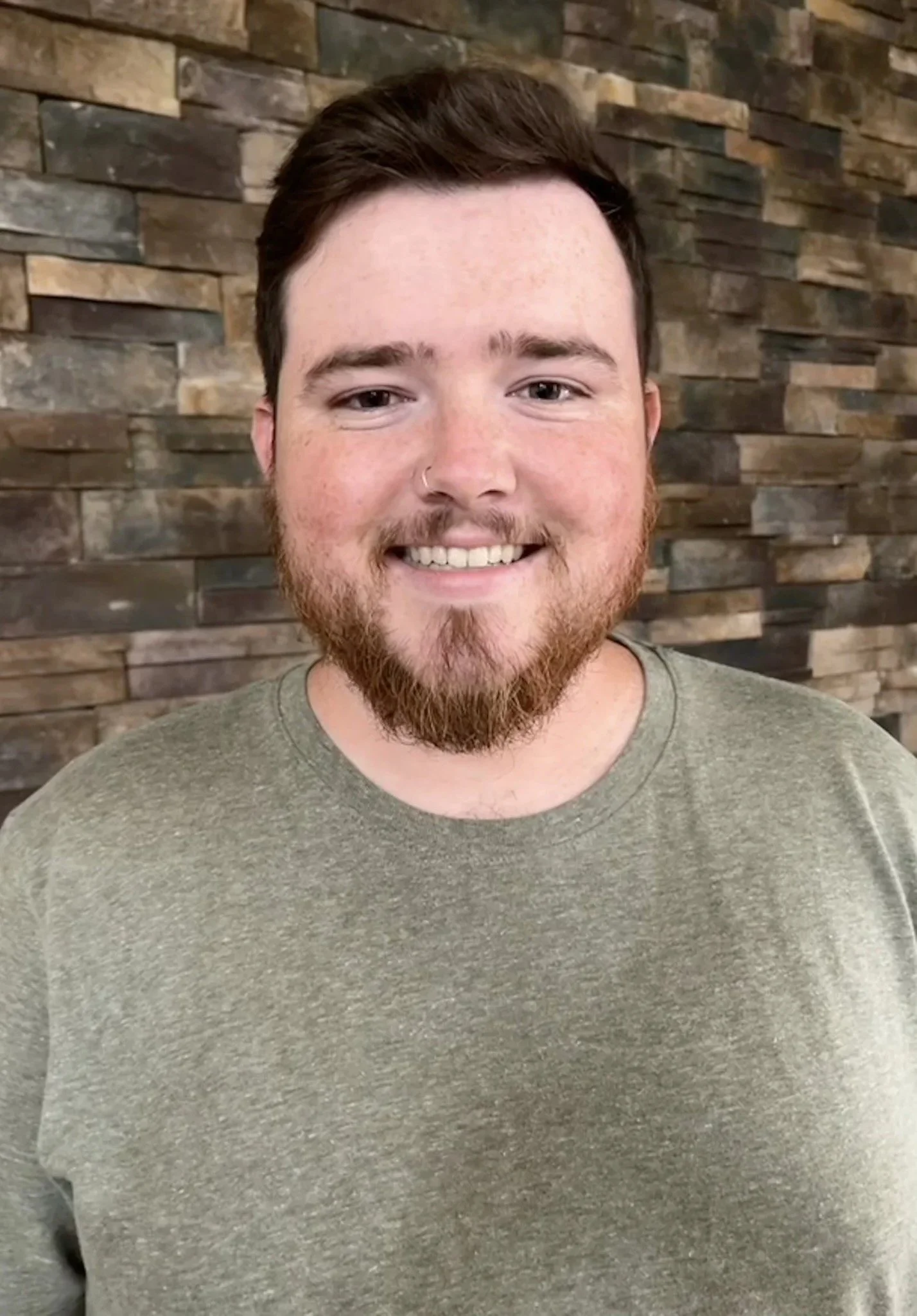 A young man with a beard and mustache smiling, wearing a green t-shirt, standing in front of a brown stone wall.