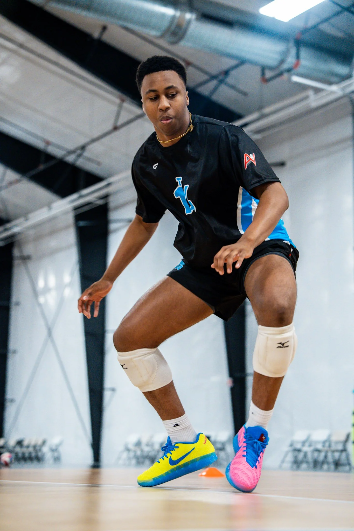 An athlete wearing a black and blue sports uniform is practicing on an indoor court. The athlete is wearing white knee pads and bright yellow and blue sneakers, with their hand reaching towards an orange cone on the floor. The background features industrial ceiling pipes and overhead lights.