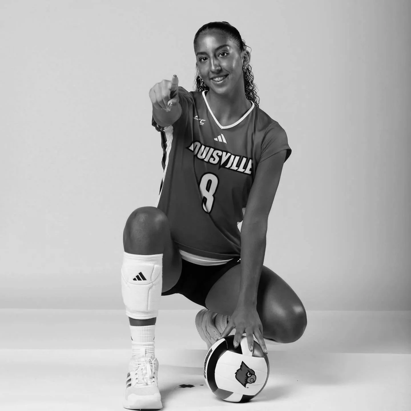 A female volleyball player in a Louisville jersey, squatting with one knee on the ground, pointing at the camera, with her volleyball and kneepad visible.
