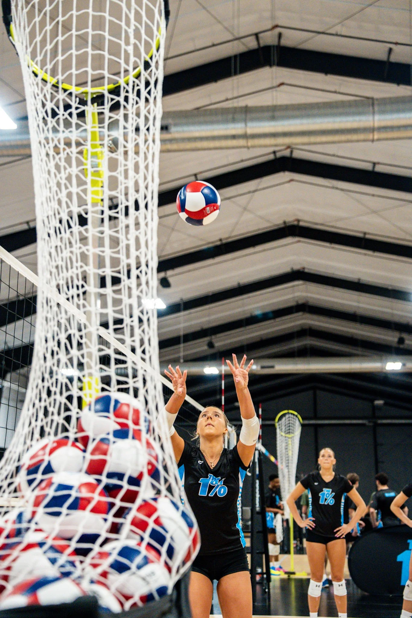 A volleyball game in action at an indoor sports facility, with a female player serving or about to hit the ball, surrounded by teammates and volleyball equipment.