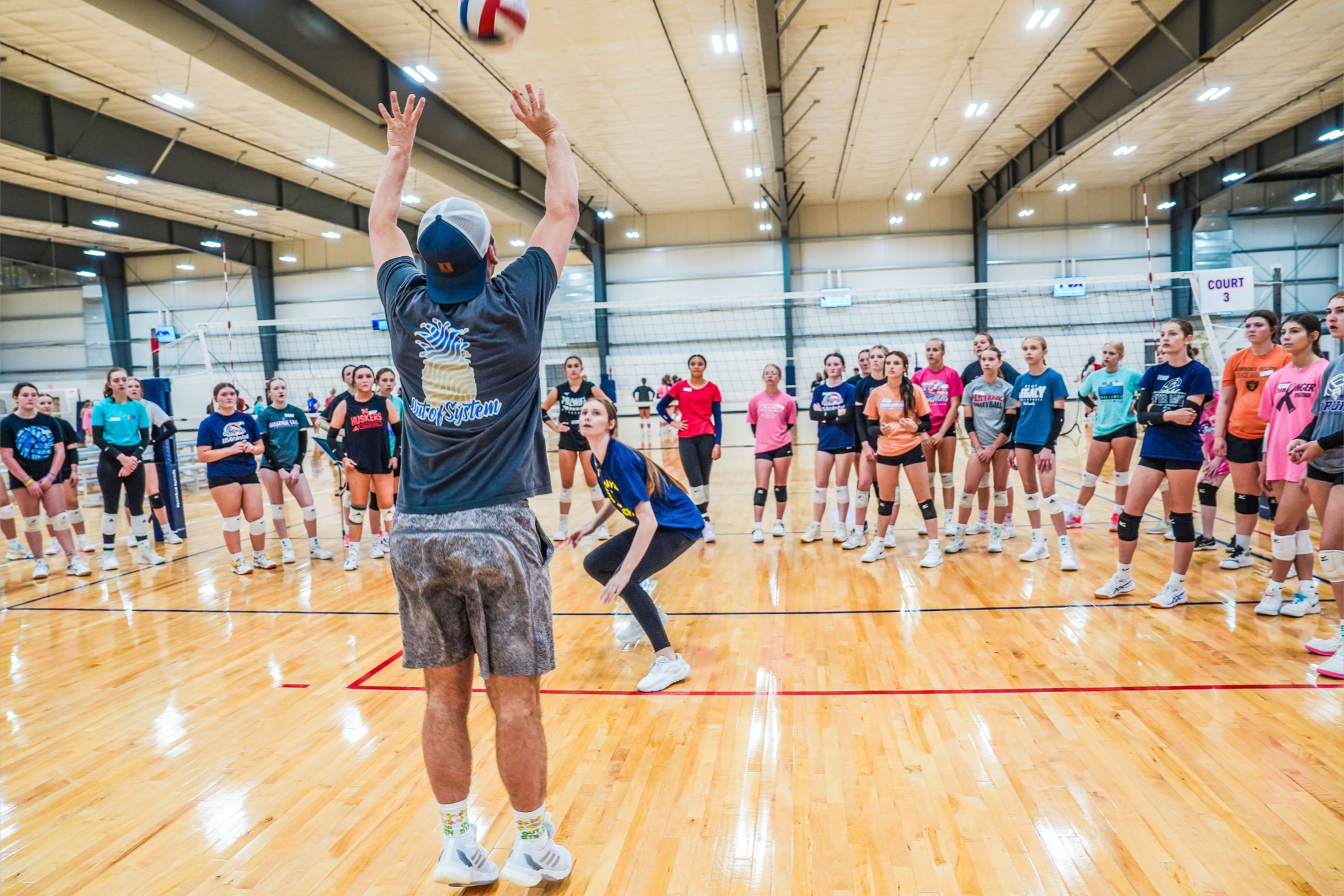 A volleyball coach in casual clothing demonstrates a volleyball technique to a group of young female players in a gymnasium.