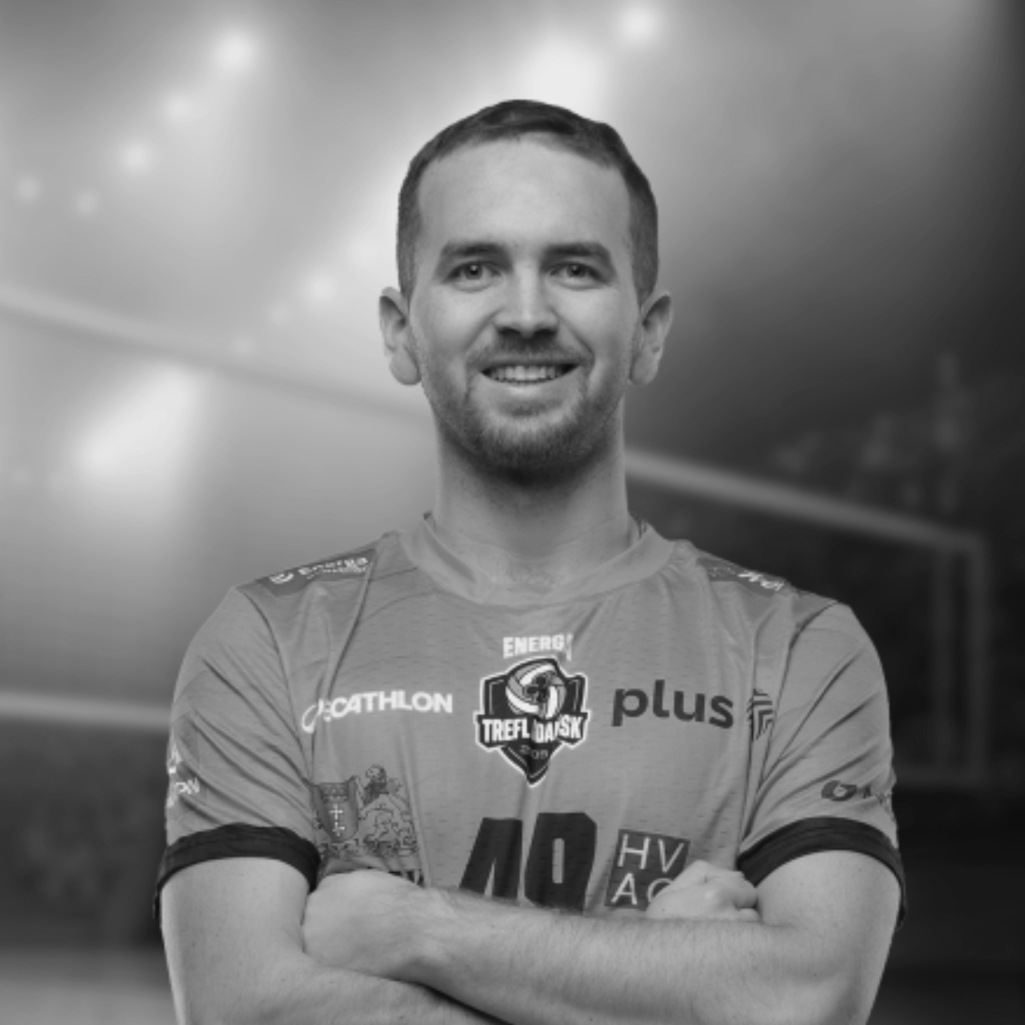 Black and white photo of a smiling Joe Worsley with short dark hair, arms crossed, wearing a sports jersey with logos and texts, against a blurred indoor background.