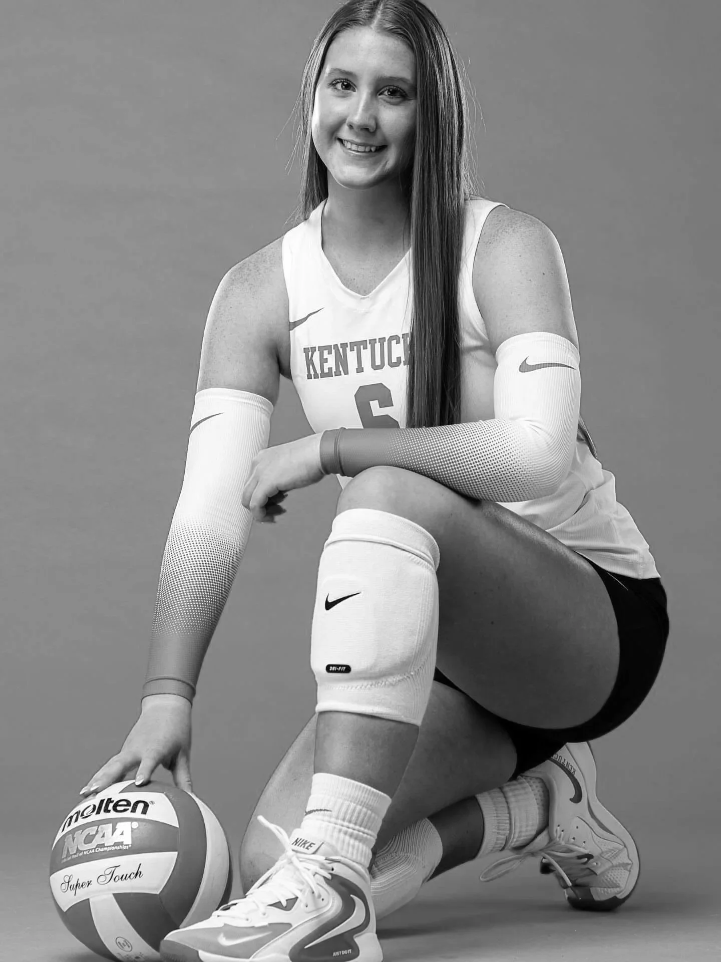 A young female volleyball player in a Kentucky jersey sitting on a stool, holding a volleyball with NCAA Super Touch logo, wearing knee-high socks and athletic shoes, smiling at the camera against a plain background.