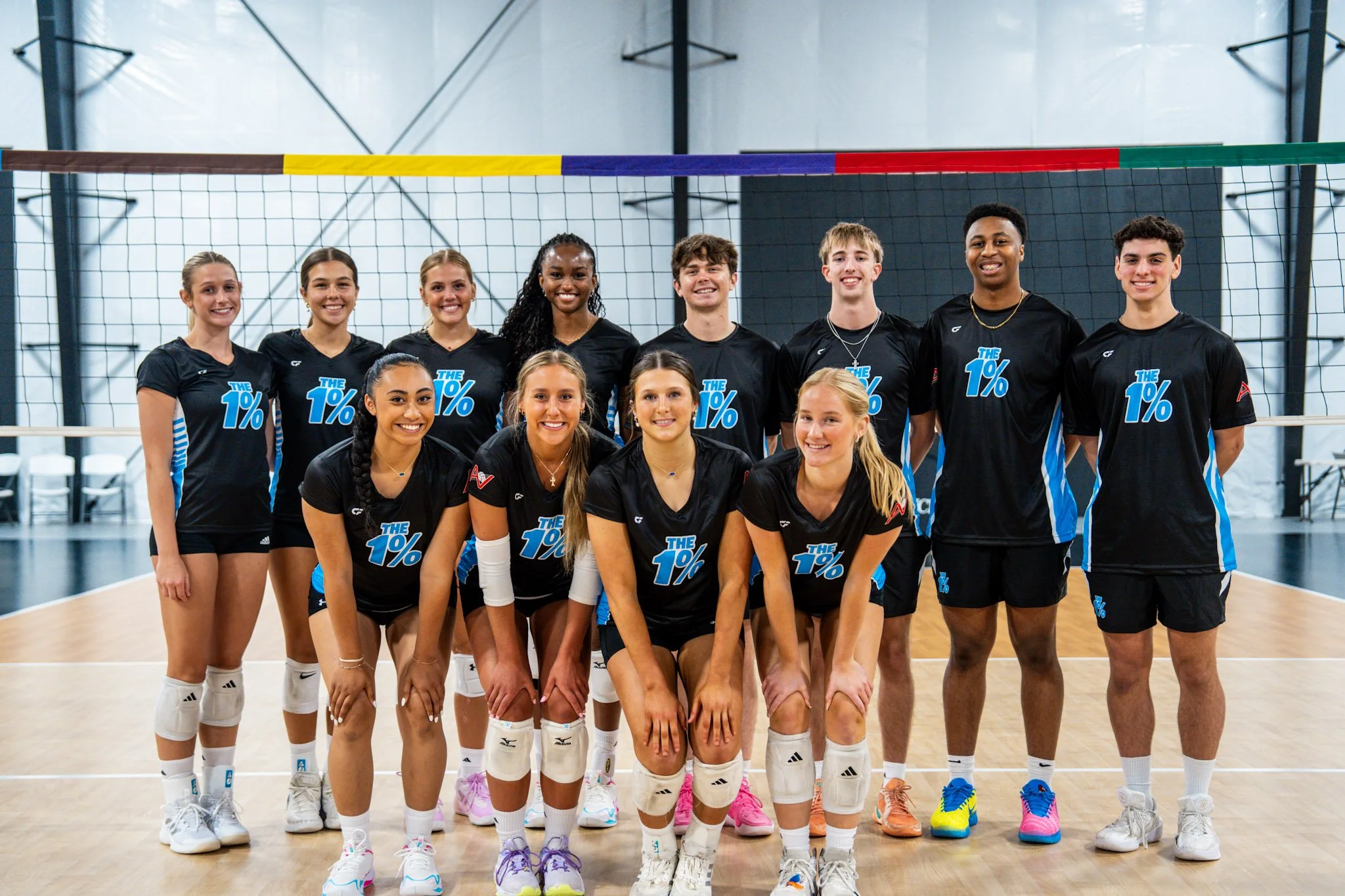 A group of young women and men volleyball players posing together on an indoor volleyball court in front of a volleyball net, smiling at the camera.