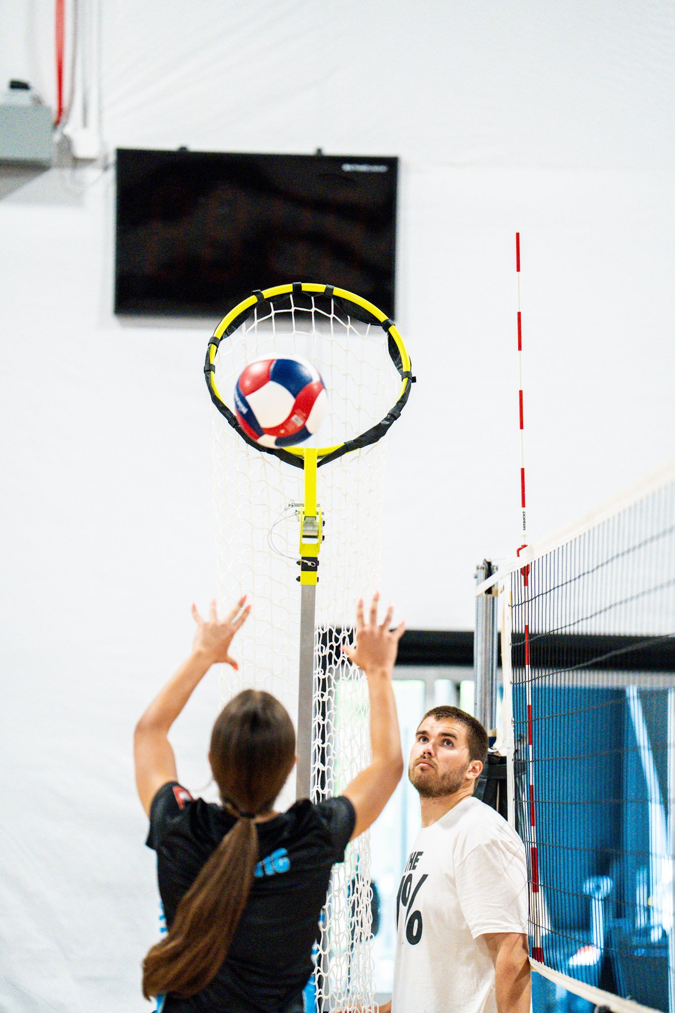 A volleyball game with a volleyball near the net, two players reaching up to block or hit, and a person watching, indoors.