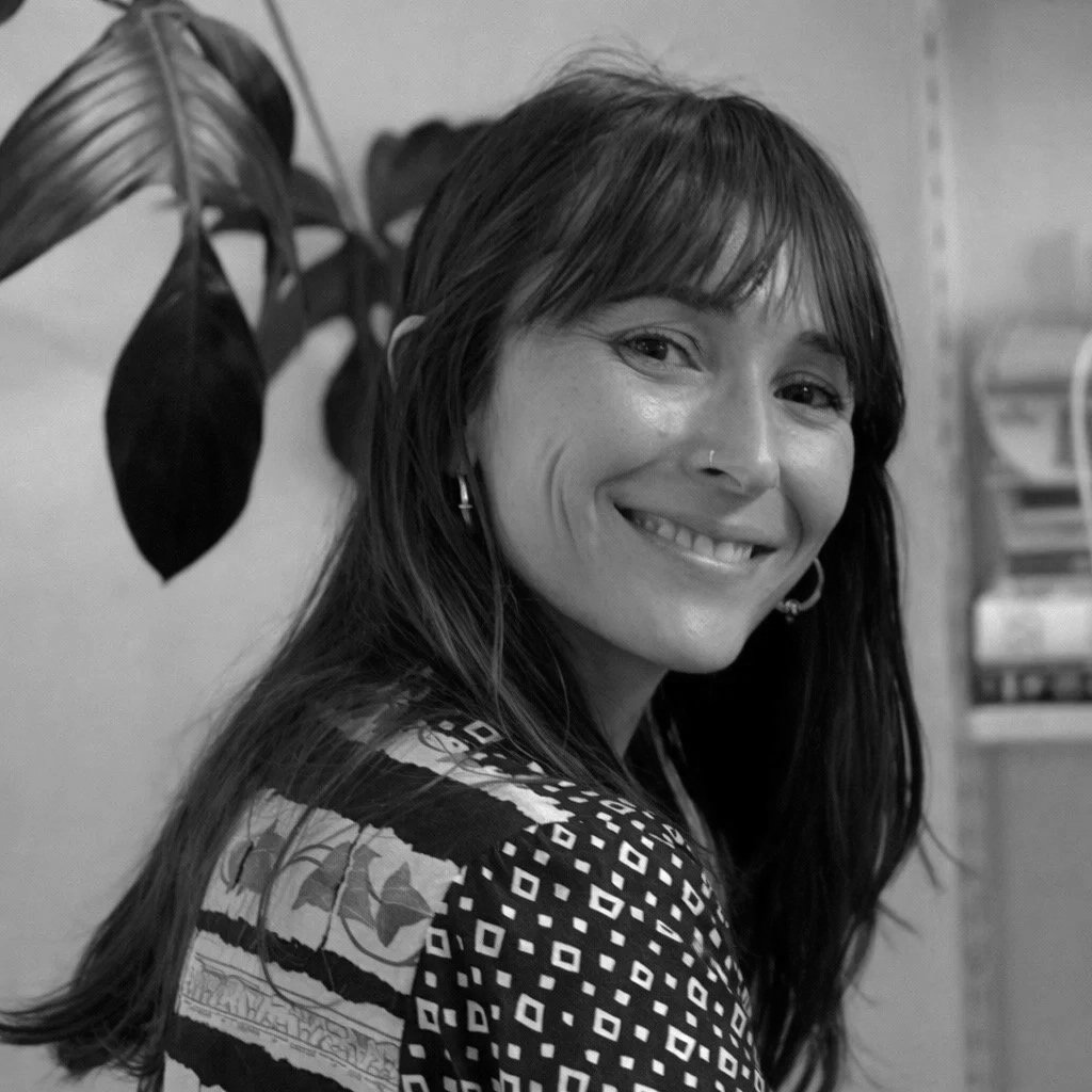 Black and white photo of a woman with long dark hair smiling at the camera, wearing earrings, a patterned top, and standing in front of a plant and some shelves.