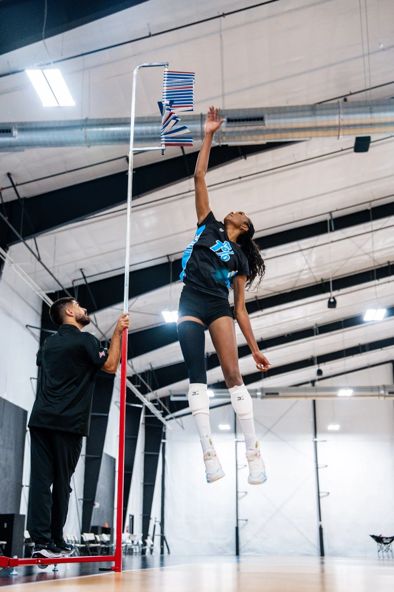 Female volleyball player jumps high to hit the ball during a practice or game in an indoor gymnasium. A coach or trainer is standing on a platform nearby, observing and possibly coaching. The gym has a high ceiling with sports lighting and structural beams.