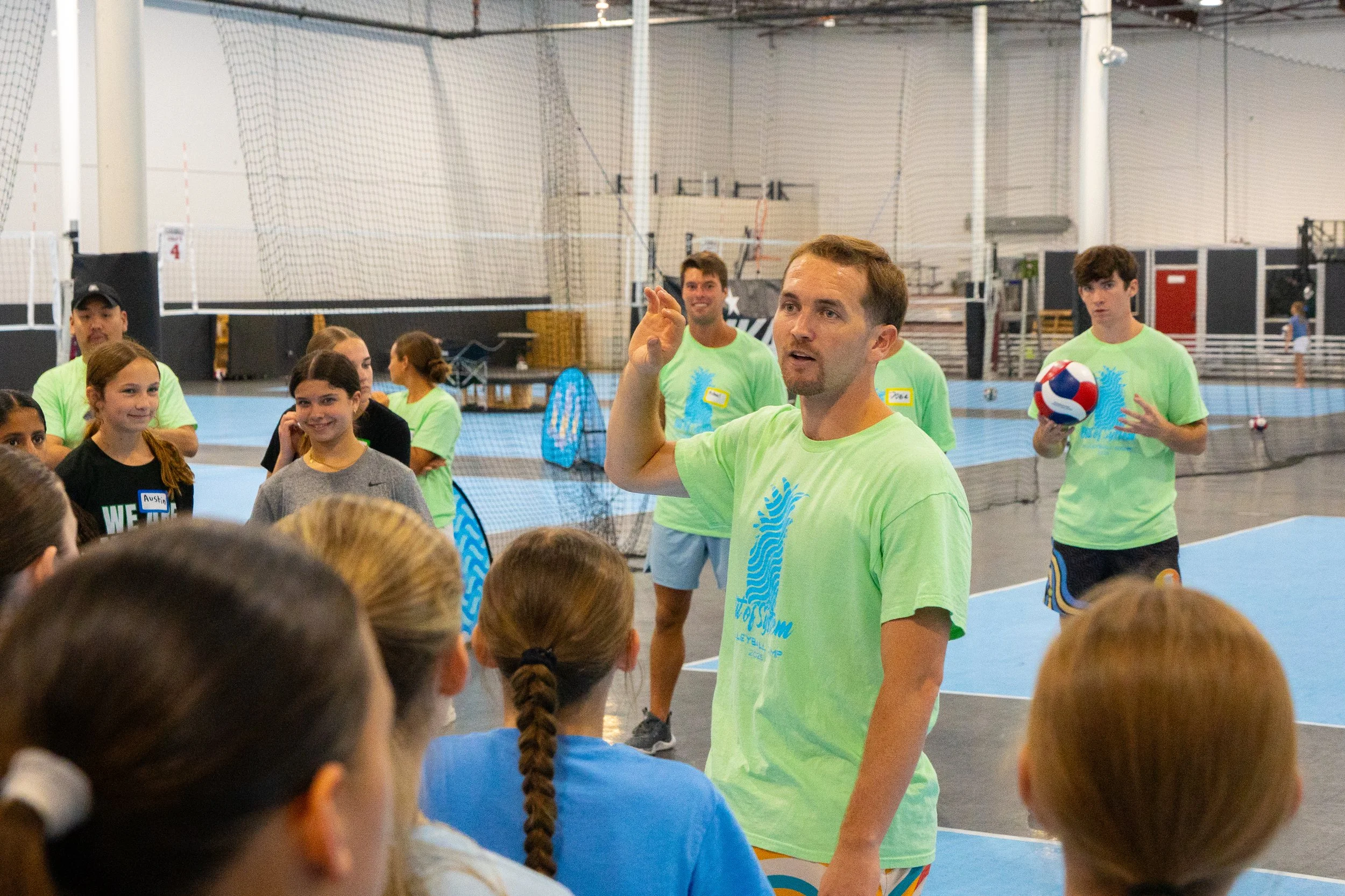 A group of children and adults in a gymnasium. An adult man is speaking to the children, who are facing him. Some people are wearing matching green t-shirts, indicating they might be part of an organized event or team. The gym has a volleyball net and sports equipment in the background.