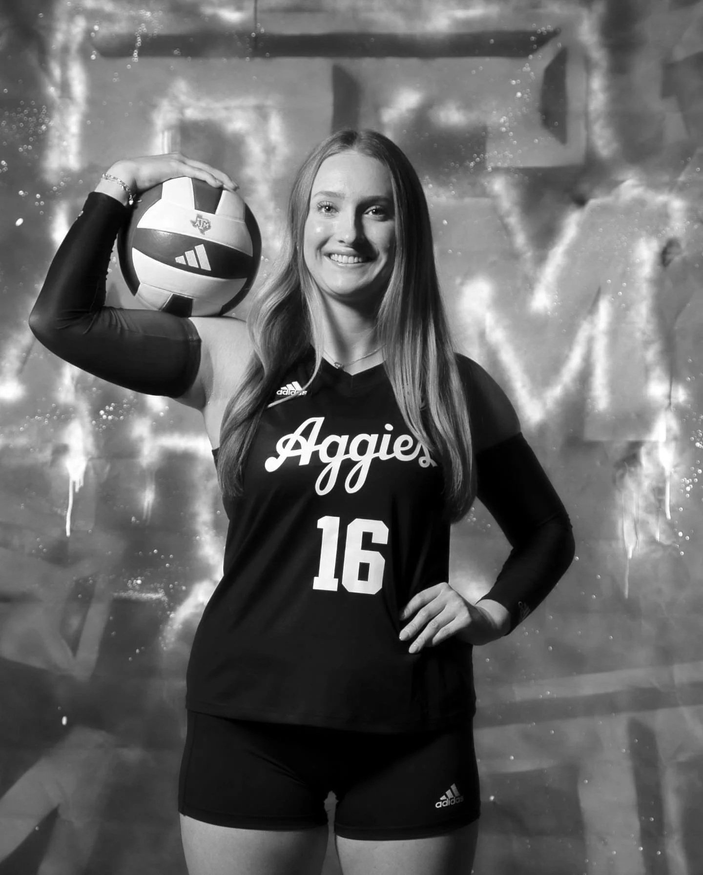 A female volleyball player stands confidently with one hand on her hip and the other holding a volleyball on her shoulder. She is smiling and wearing a black sports uniform with the word 'Aggies' and the number 16. The background appears to be an indoor volleyball court with a reflective surface.
