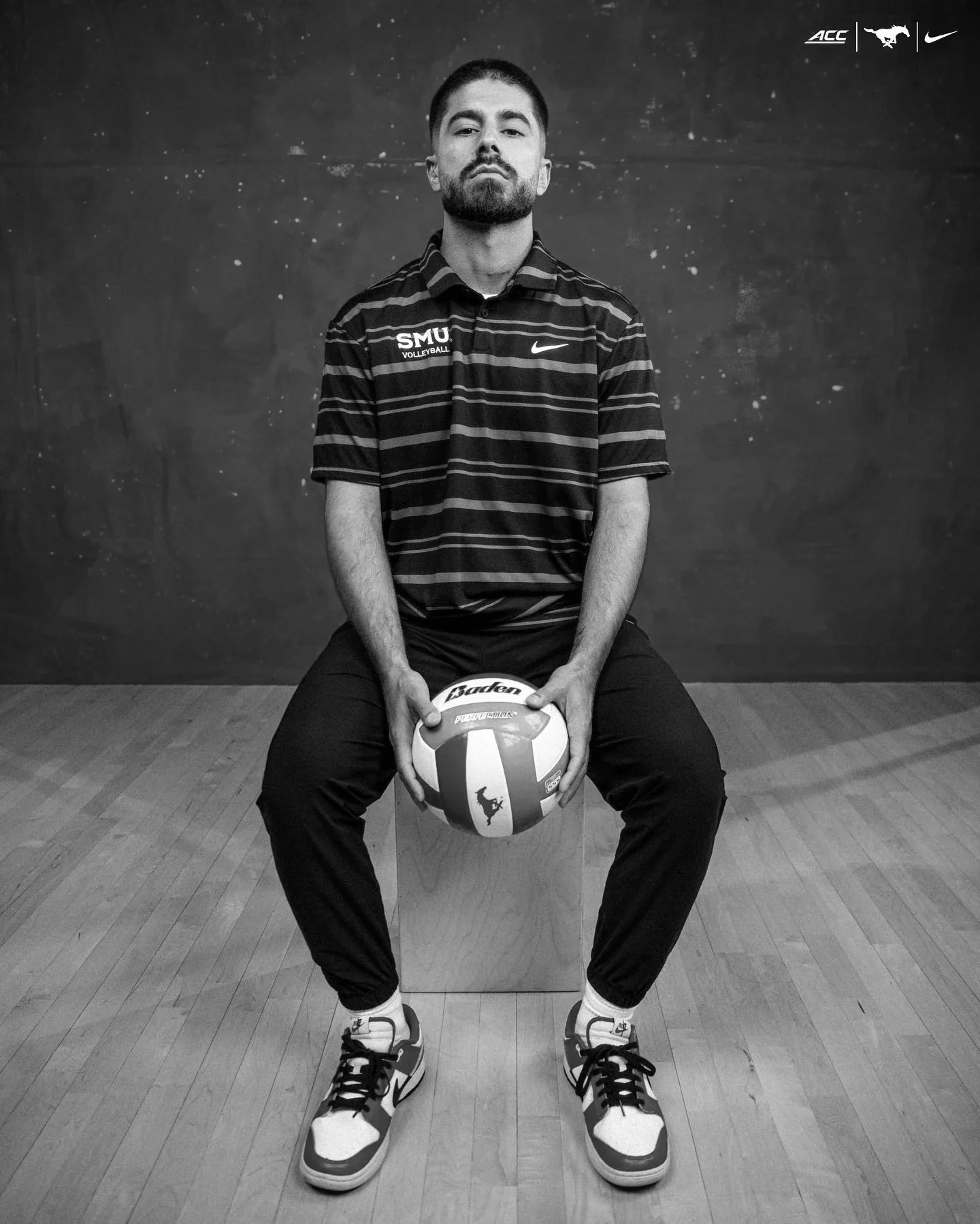 Black and white image of a man sitting on a bench, holding a volleyball with both hands, in a sports setting with a wooden floor and a plain wall. The man has short hair, a beard, and is wearing a collared shirt with the word 'SMU Volleyball' and a Nike logo, along with sports shoes. Logos of ACC, Mustang, and Nike are visible in the top right corner.