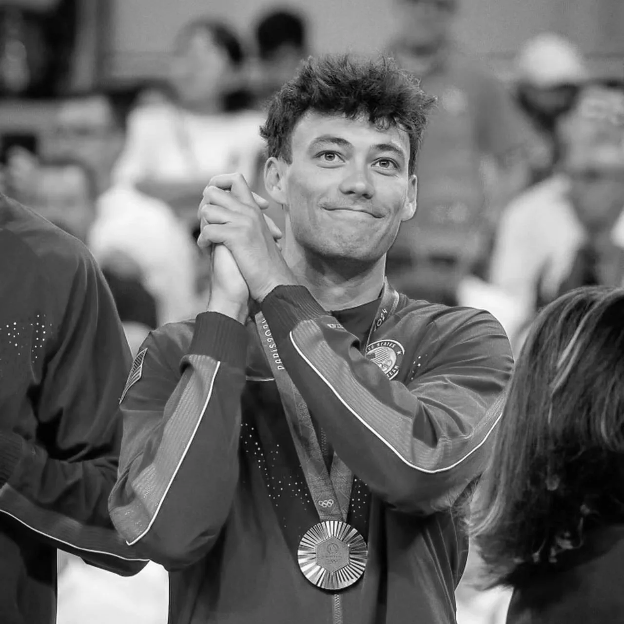 Micah Ma'a wearing a jacket with an American flag patch, smiling while holding his hands together near his face. He is wearing a medal around his neck,  at a sporting or award event, with spectators or other attendees visible in the background.