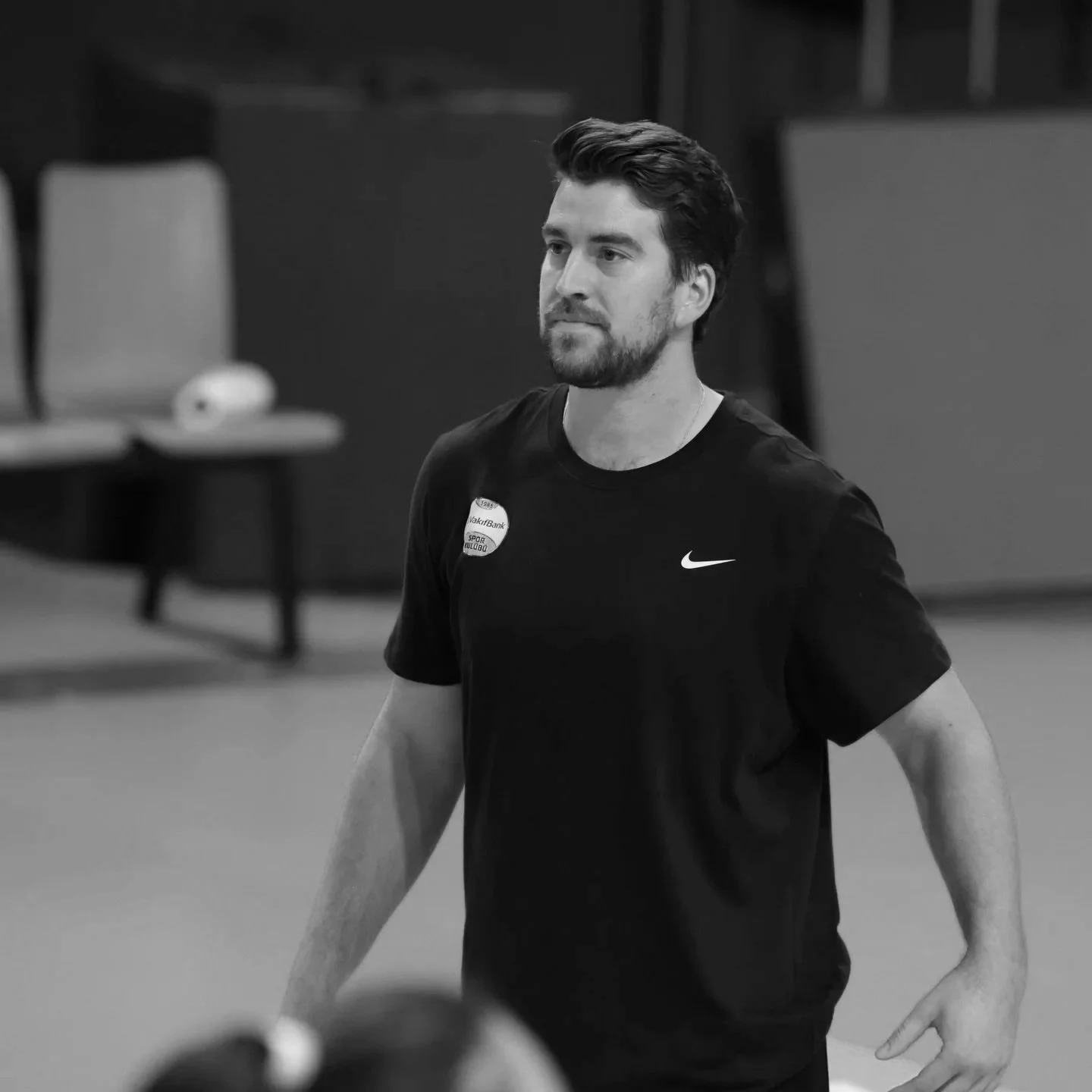 A young man with dark hair and a beard is standing in an indoor sports facility. He is wearing a black Nike T-shirt with a badge on it. Chairs and a table with some objects are in the background.