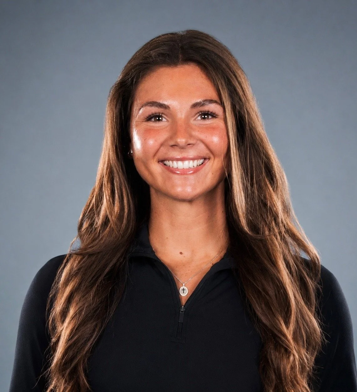 A young woman with long wavy brown hair, smiling, wearing a black zip-up athletic top and a cross necklace, standing against a neutral background.
