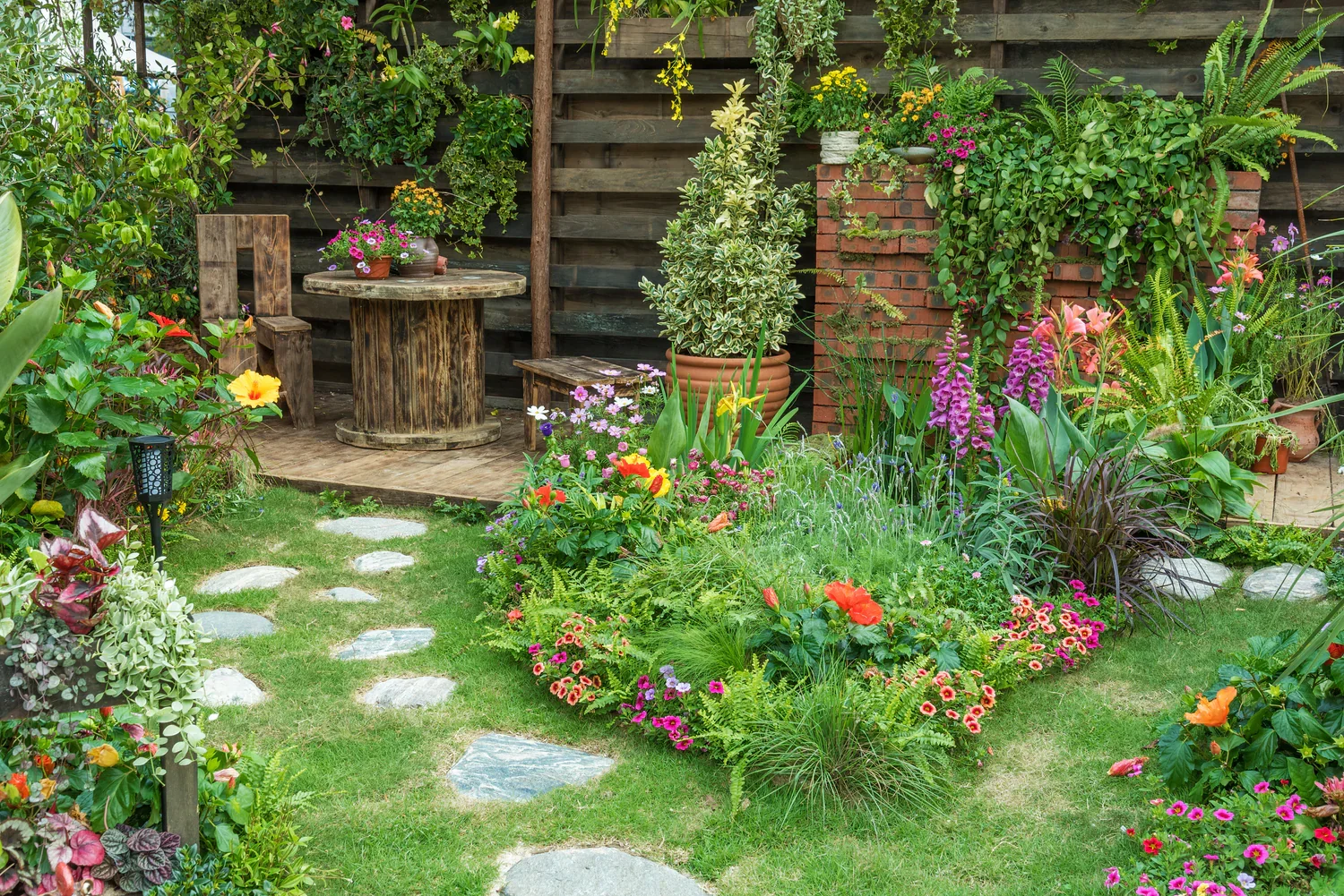A colorful garden with a variety of blooming flowers, a grassy lawn with stepping stones, potted plants, and a small wooden patio with a round table and two benches, all enclosed by a wooden fence.