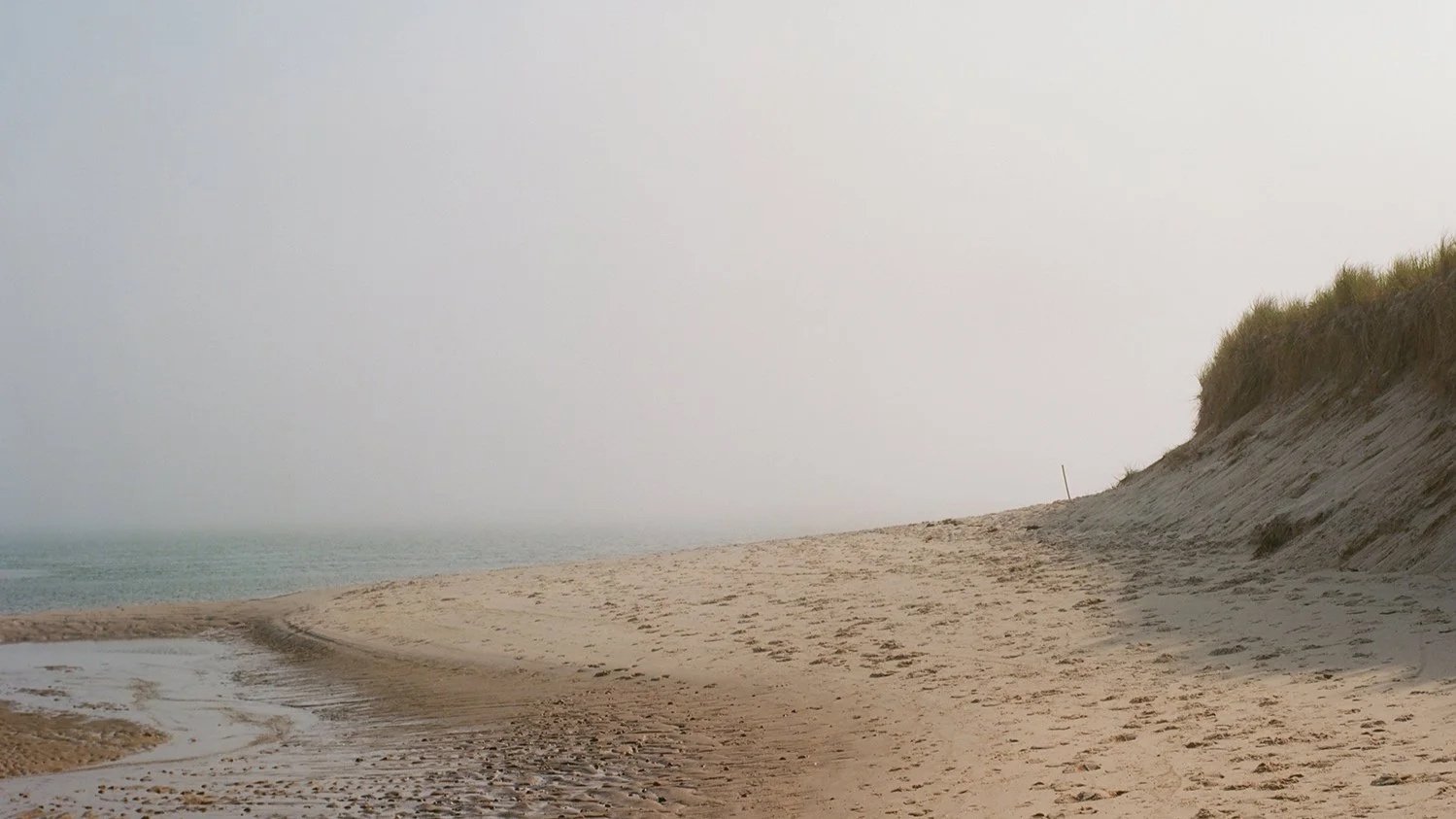 Empty sandy beach with a grassy dune on the right and a hazy sky over the ocean in the distance.