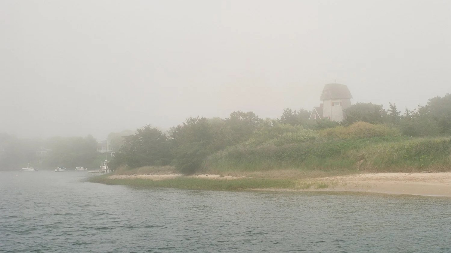 A misty scene of a river with a sandy shoreline and lush green trees on the riverbank. In the background, a lighthouse or tower is visible through the fog.
