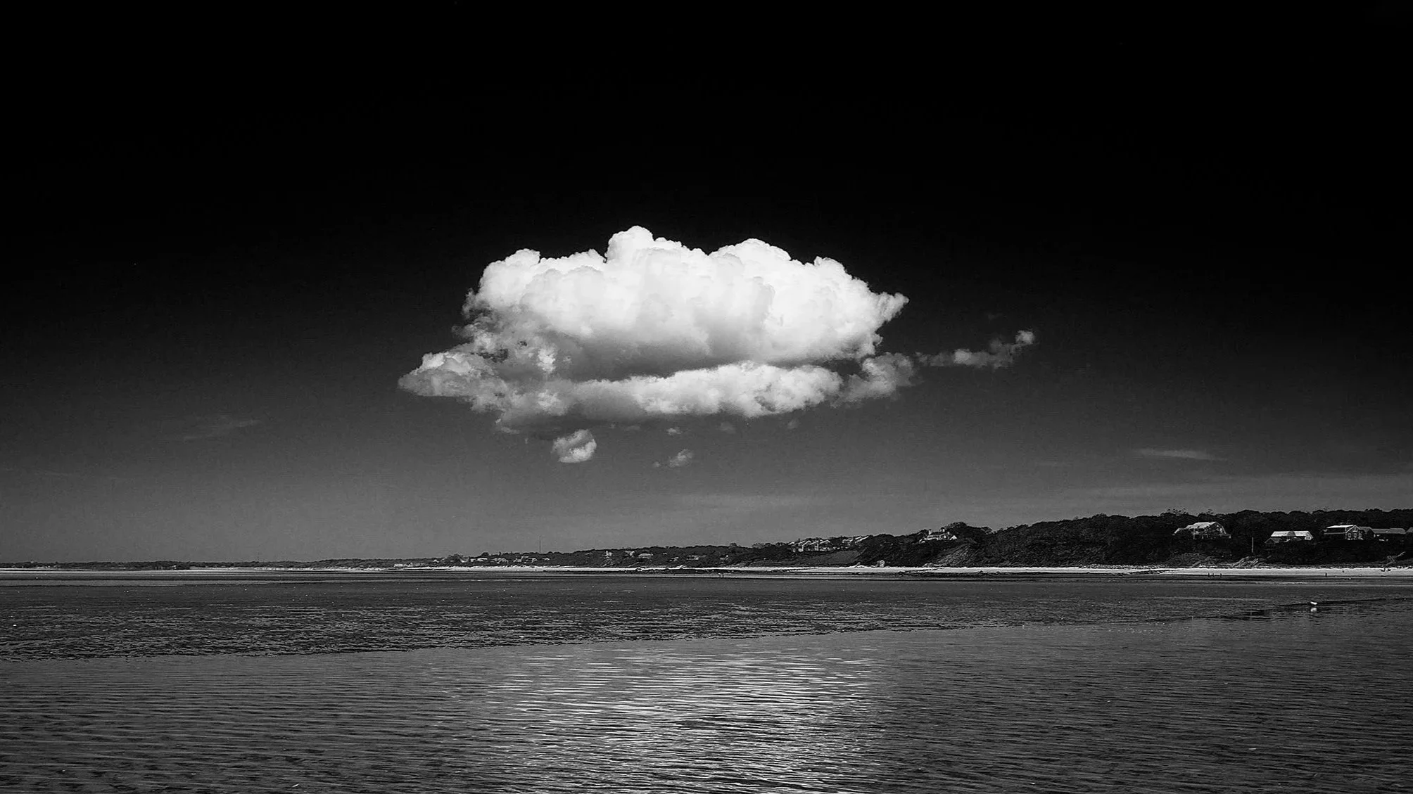 Black and white photograph of a lone cloud out on Brewster Flats, Cape Cod with reflection in the ocean and scenery in the background.