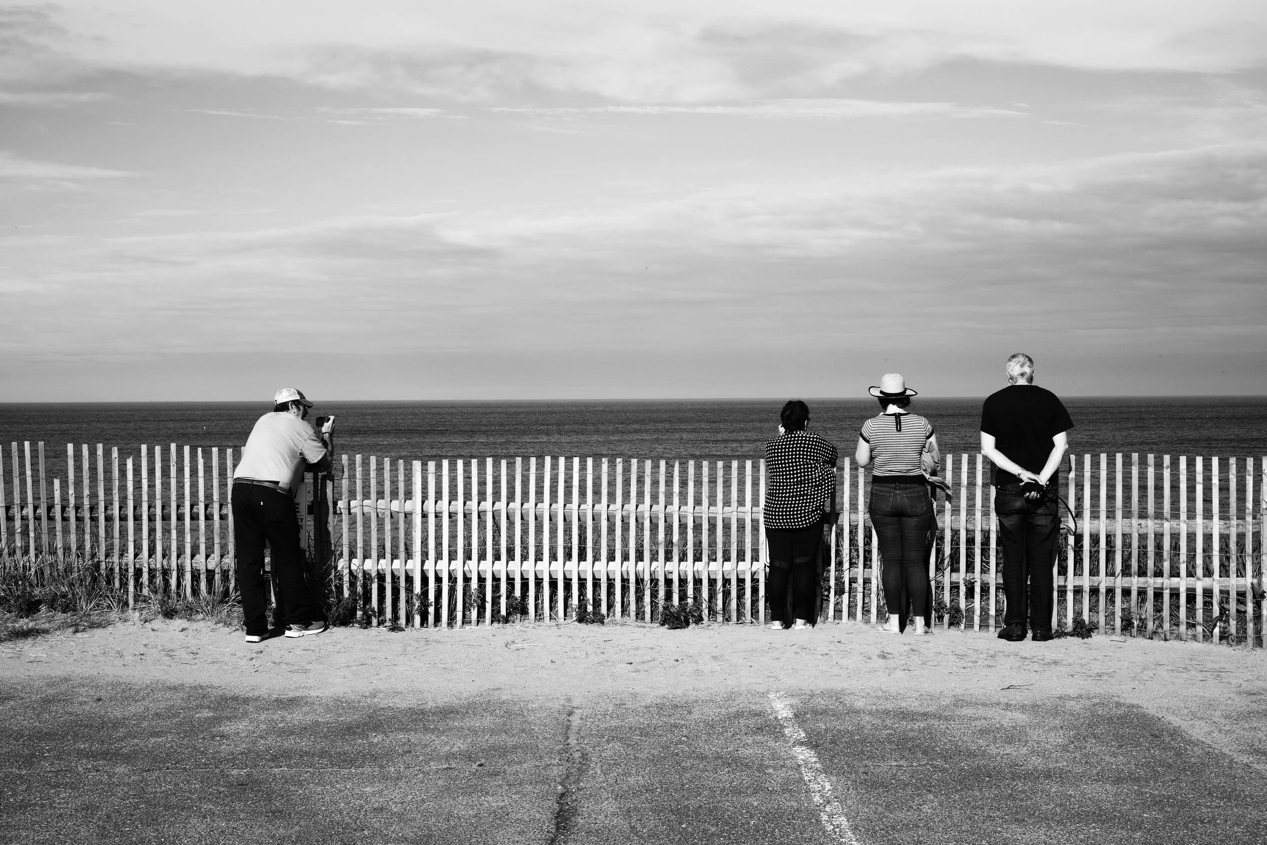 Black and white photograph of four people leaning on a wooden picket fence and looking out to sea on National Sea Shore Cape Cod