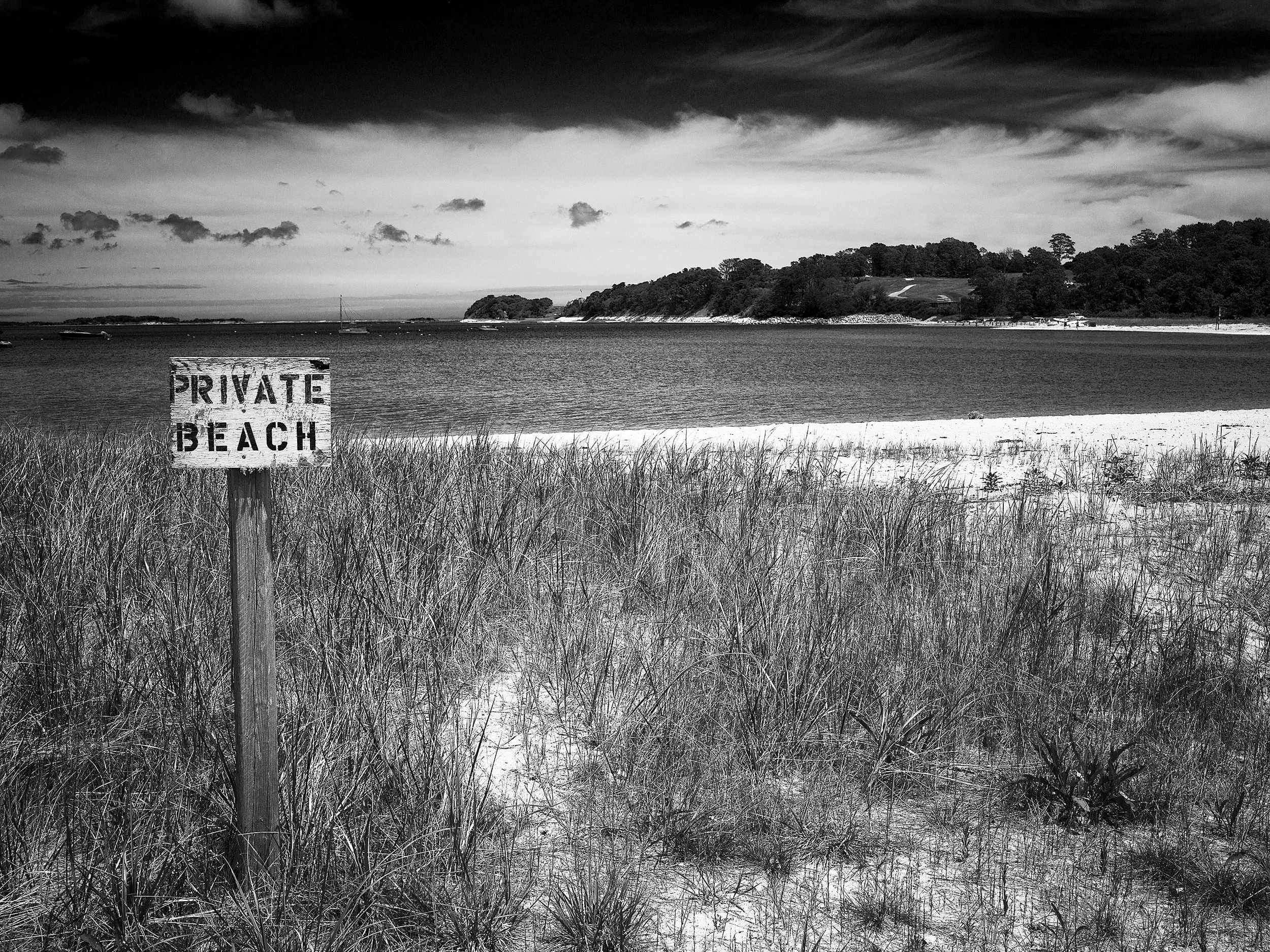 A black-and-white image of a beach with a grassy foreground, water, and a distant island or shoreline with trees. There is a sign in the grass that reads 'PRIVATE BEACH'.