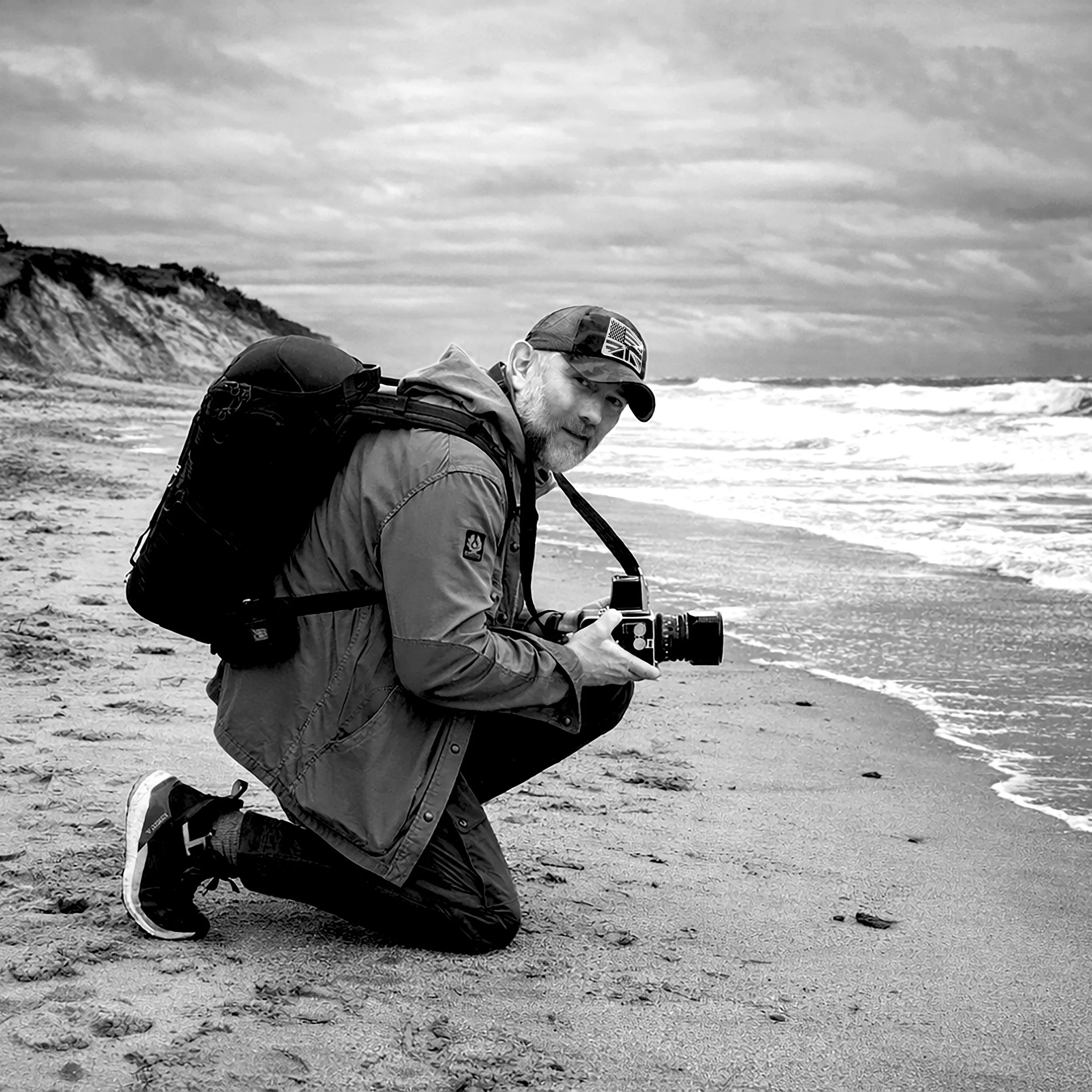 A man with a beard kneeling on a sandy beach, holding a camera, with cliffs and the sea in the background, and wearing a cap with the Union Jack.
