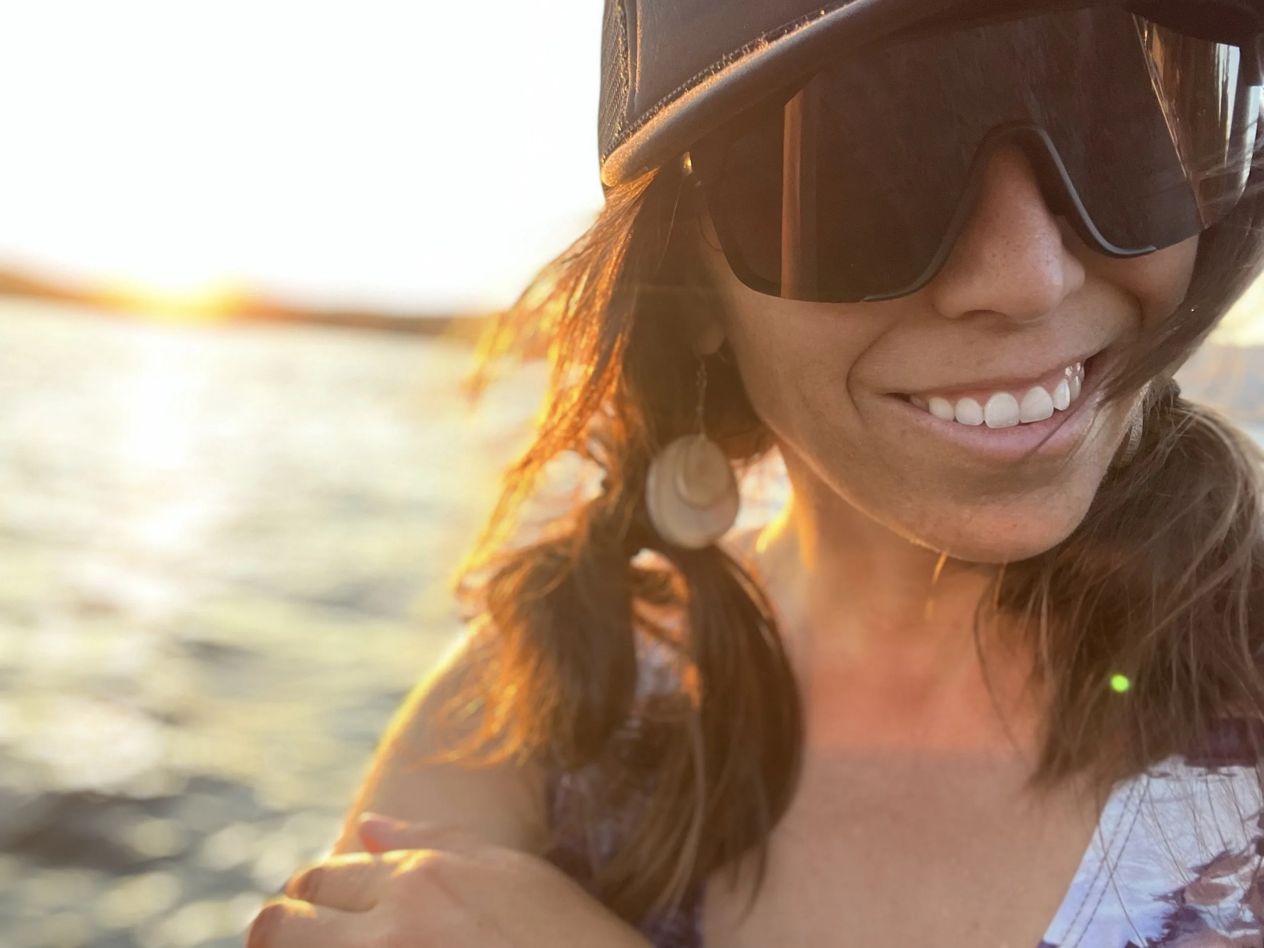 Close-up of a smiling woman wearing sunglasses and a hat, with a sunset over water in the background.