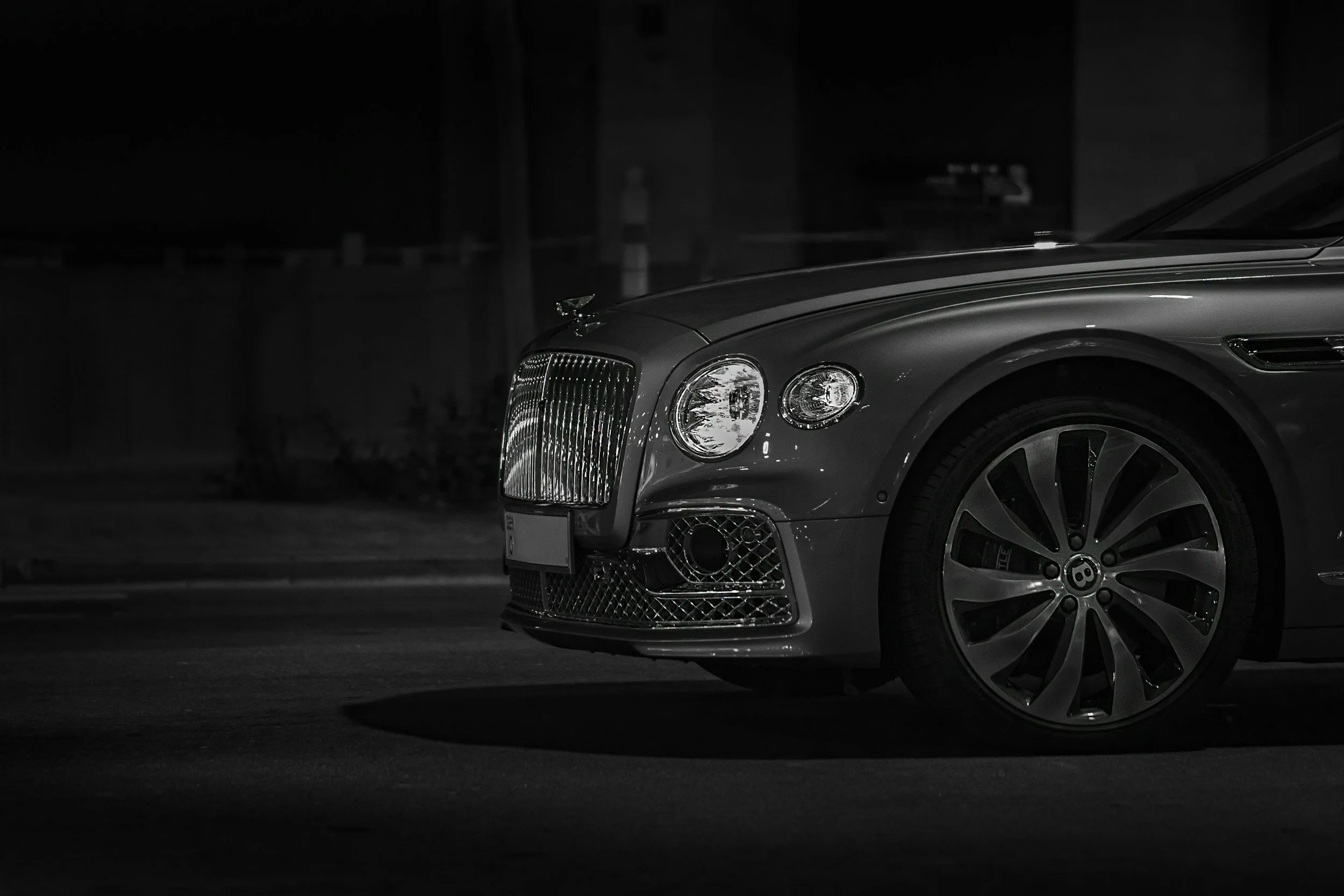 A close-up of the front part of a luxury car, showing the grille, headlights, and right front wheel, parked at night with shadows on the ground.