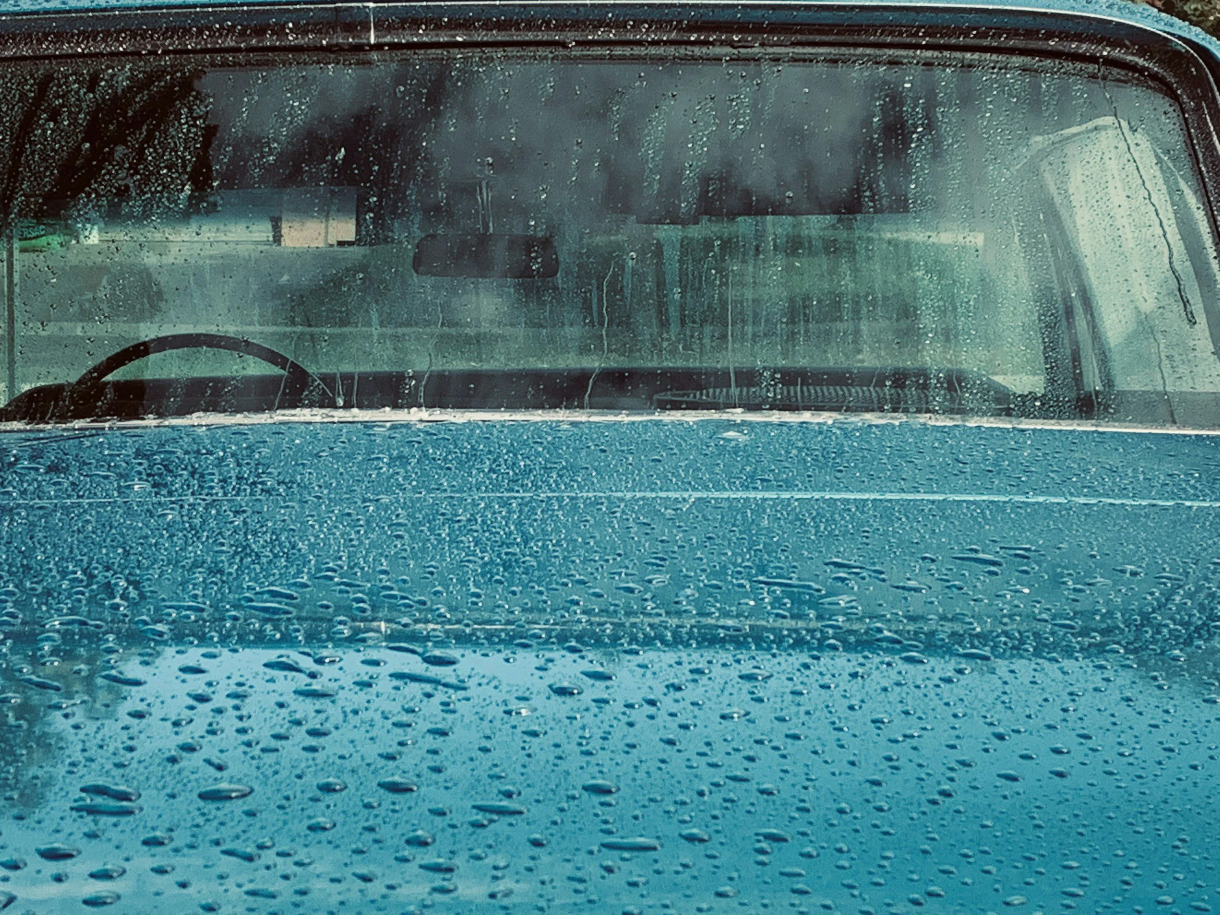 Close-up view of the front of a blue car's hood and windshield with raindrops on the surface, through which the interior of the vehicle and the steering wheel are visible in a rainy setting.