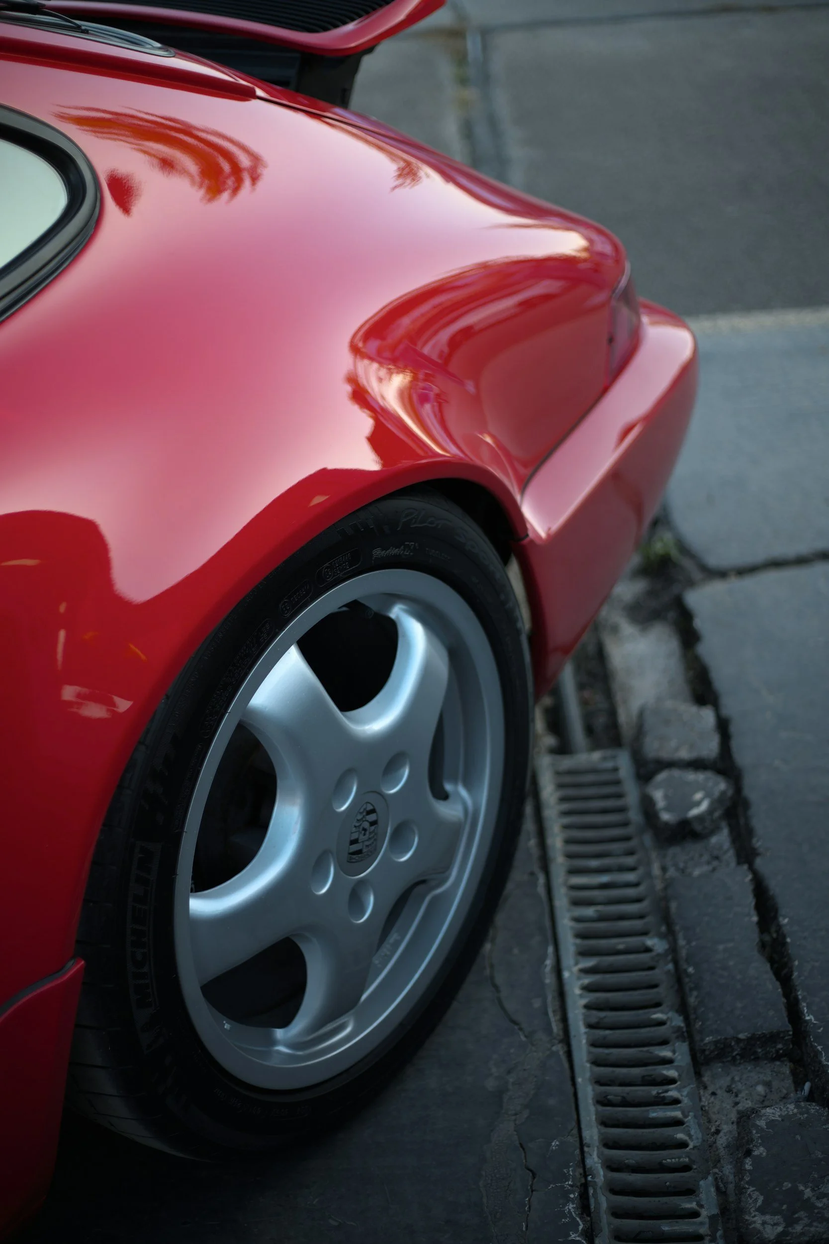 Close-up of the rear section of a red sports car showing a portion of the wheel and tire, parked on a sidewalk.