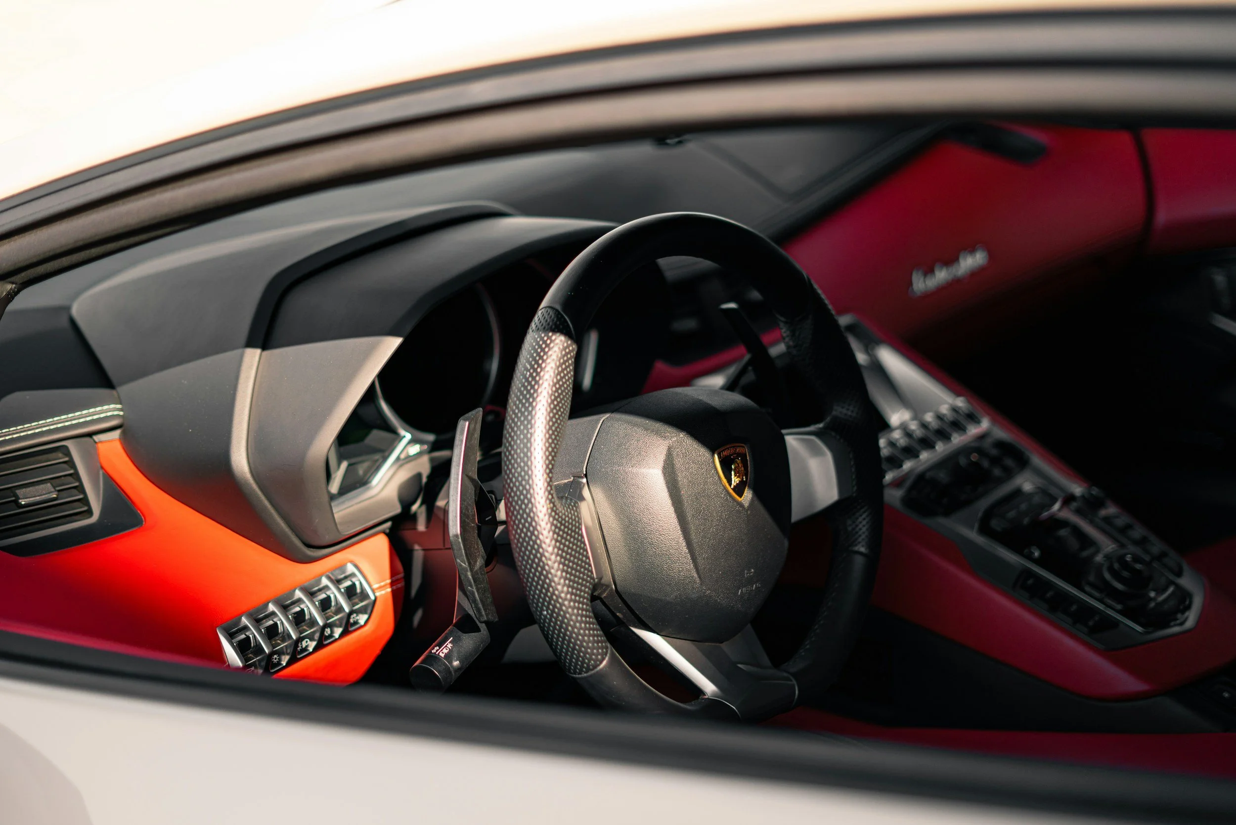 Inside view of a luxury sports car with a black and red interior, and a Lamborghini emblem on the steering wheel.