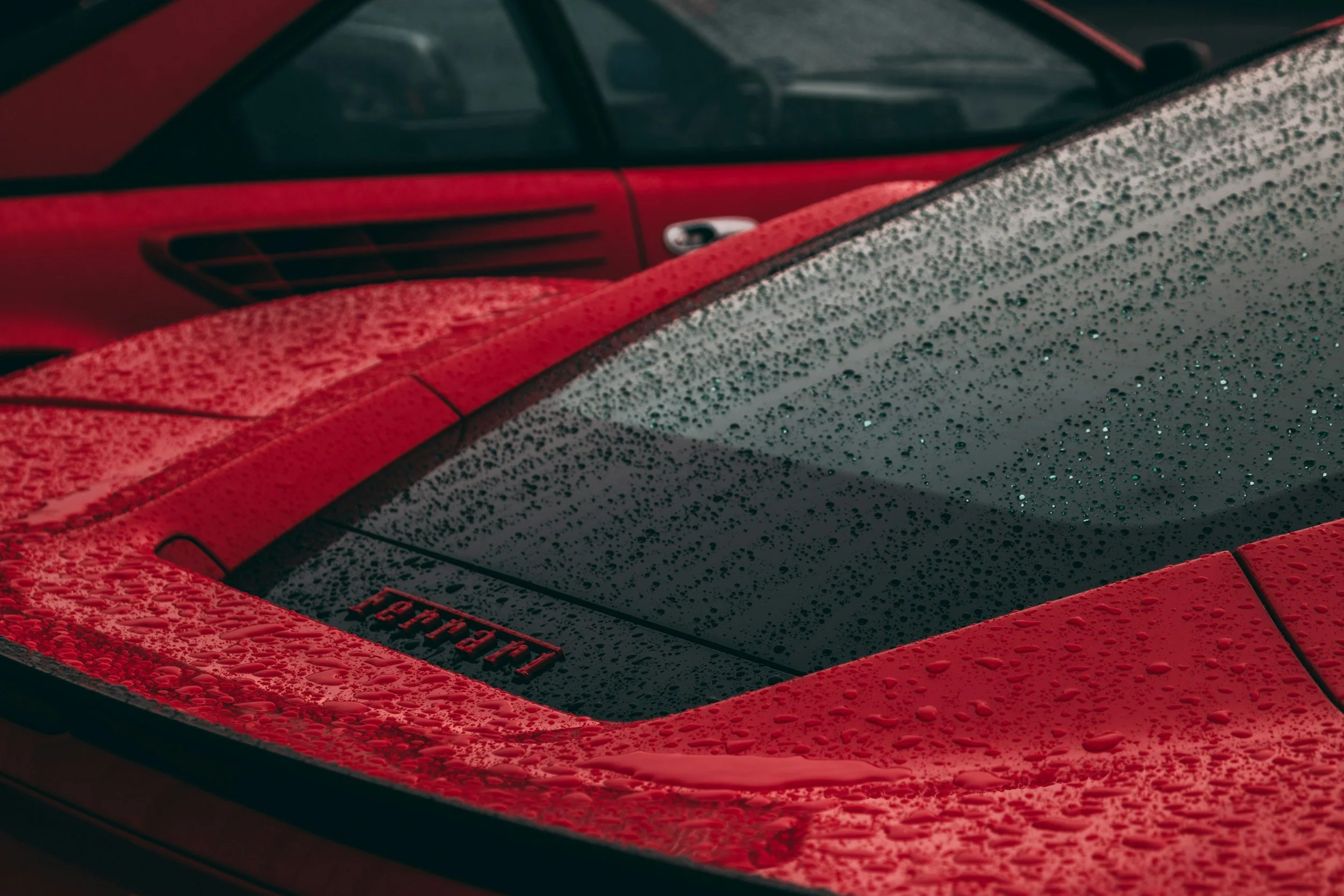 Close-up of a red Ferrari sports car with water droplets on the rear window and body, showing the Ferrari logo.