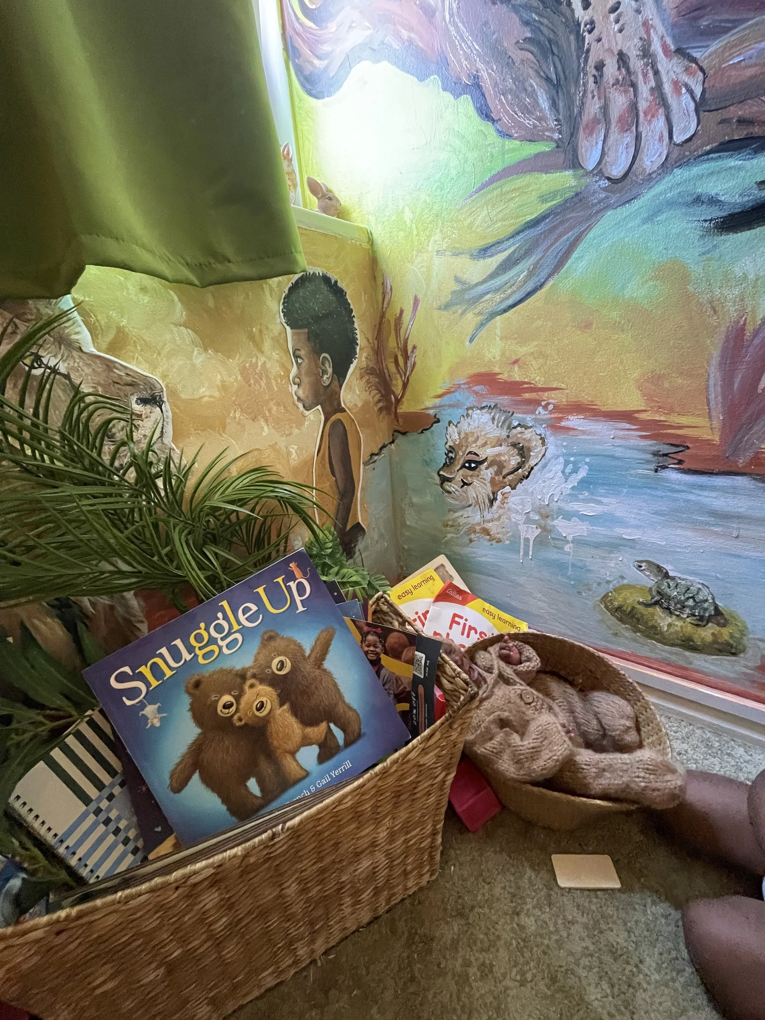 A colorful wall mural of a young child with dark skin and short curly hair, and a lion cub, in a natural setting with water, a crocodile, and a bird, with a basket of children's books and toys in the foreground.