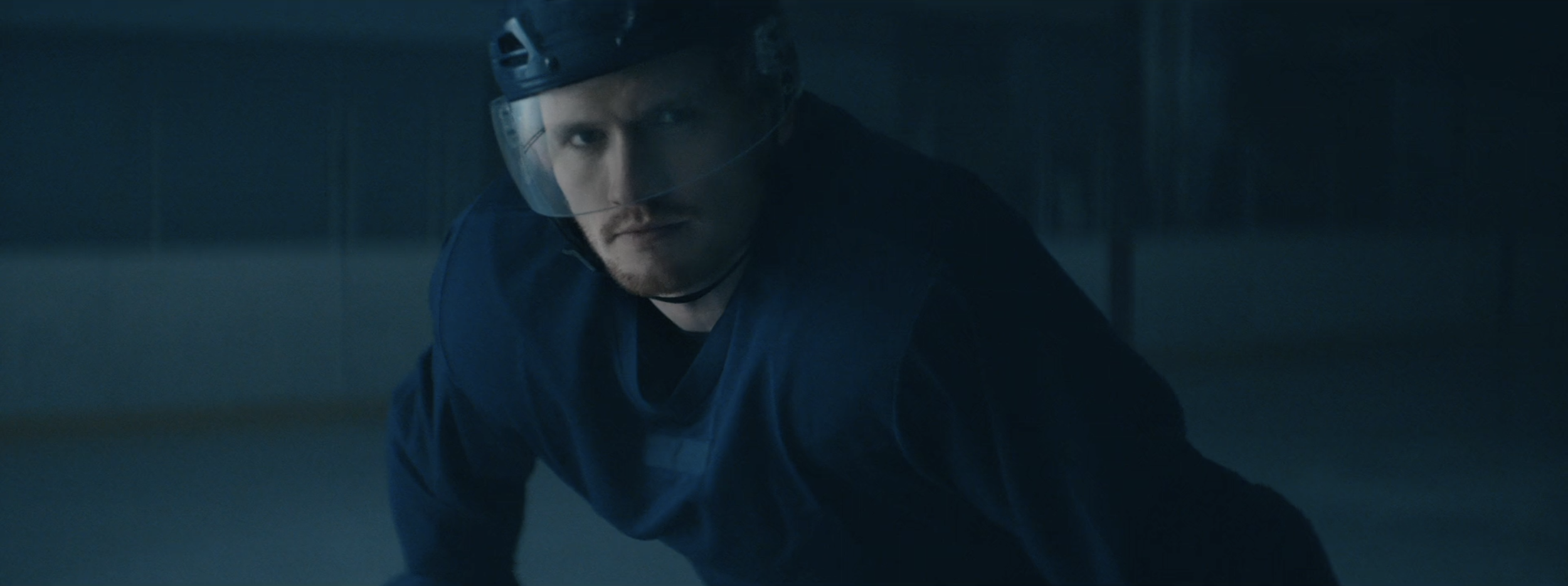A man wearing a helmet and dark clothing is crouched down on the ground in a dimly lit room, looking intently forward.