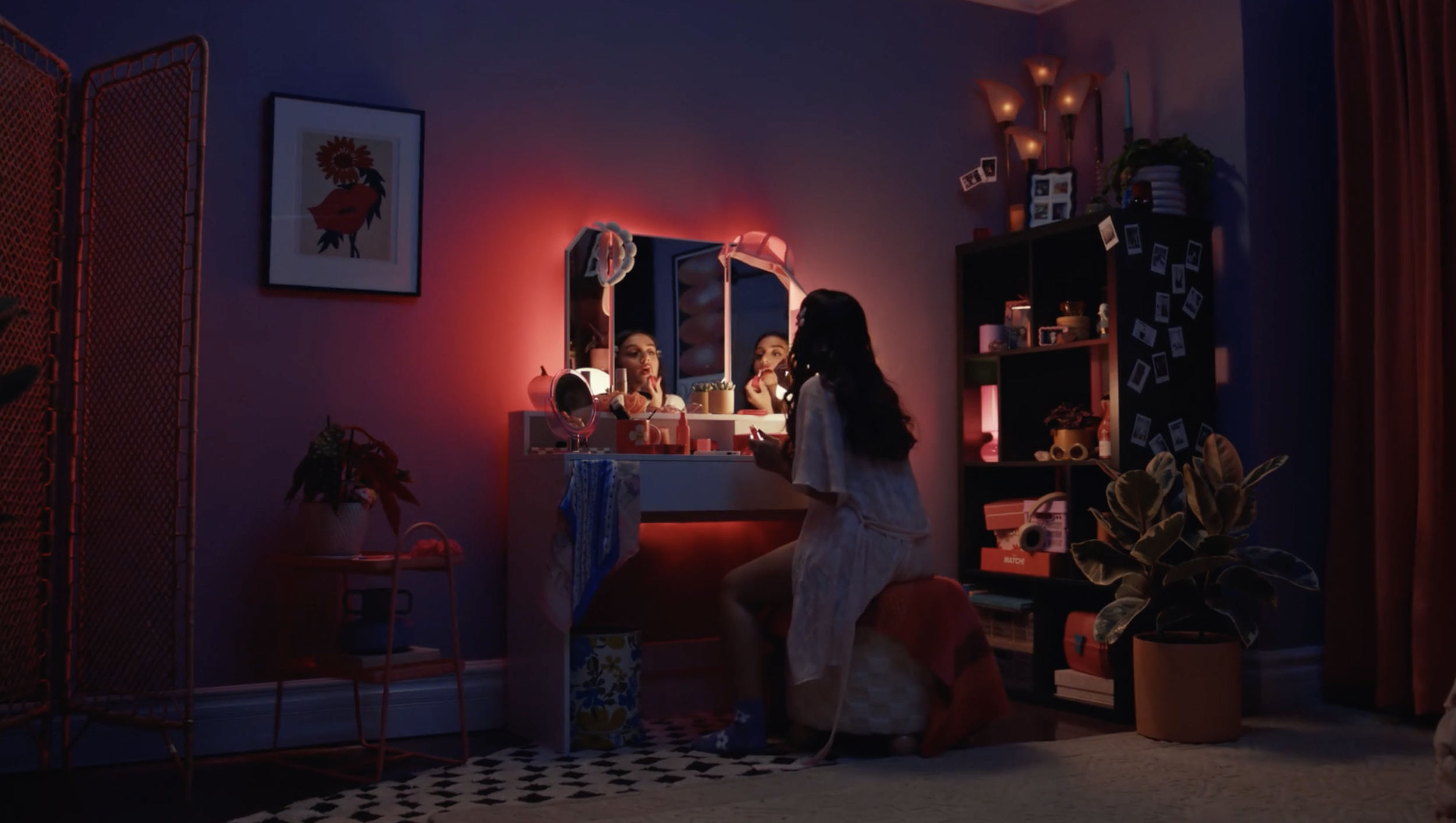 A woman sitting at a vanity table in a dimly lit room, applying makeup while looking into a mirror. The room has colorful, warm lighting, a black bookshelf, potted plants, and decorative items, with photos on the side of the bookshelf.