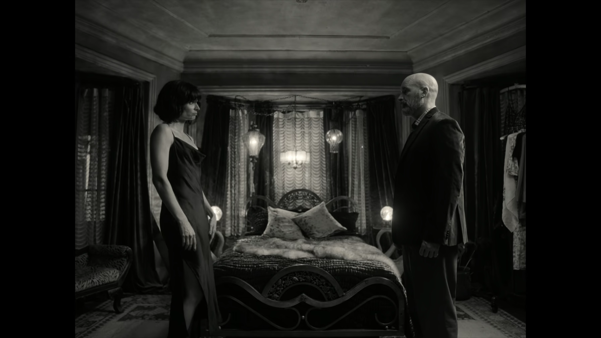 Black and white image of a woman in a nightgown and a man in a suit standing in a dimly lit bedroom facing each other, with a bed and decorative curtains in the background.