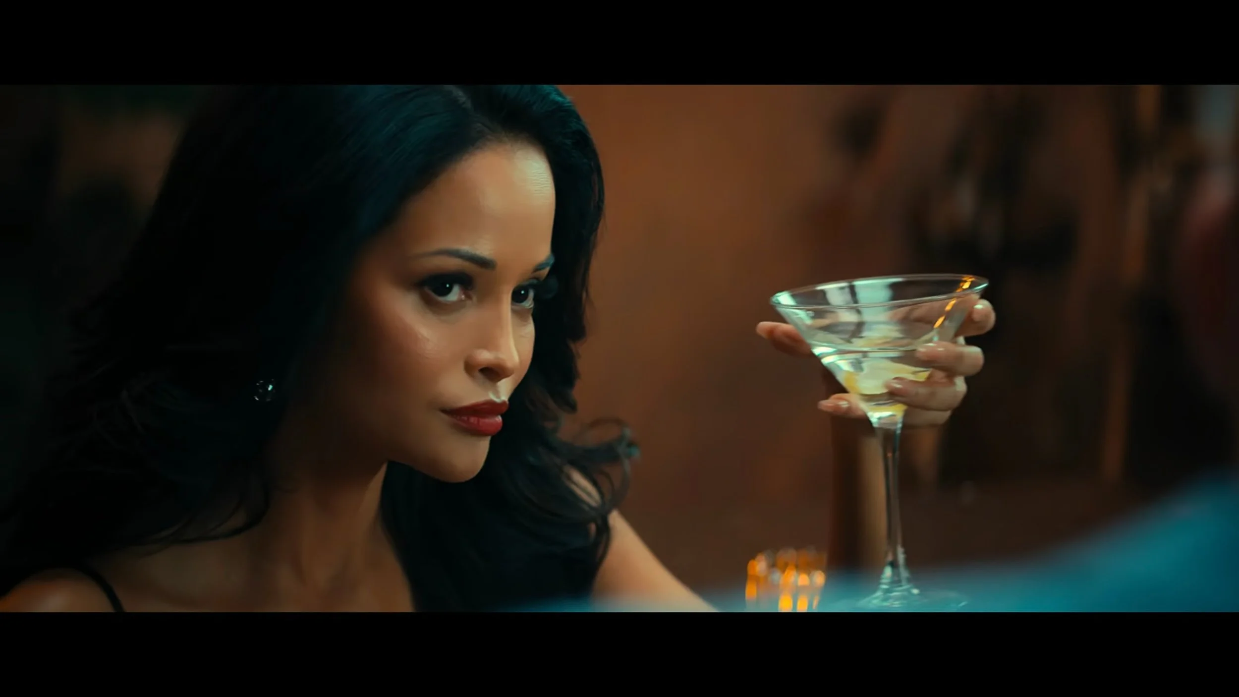 A woman with long dark hair and red lipstick sitting at a bar, holding a martini glass filled with a clear drink, looking intently to her left.
