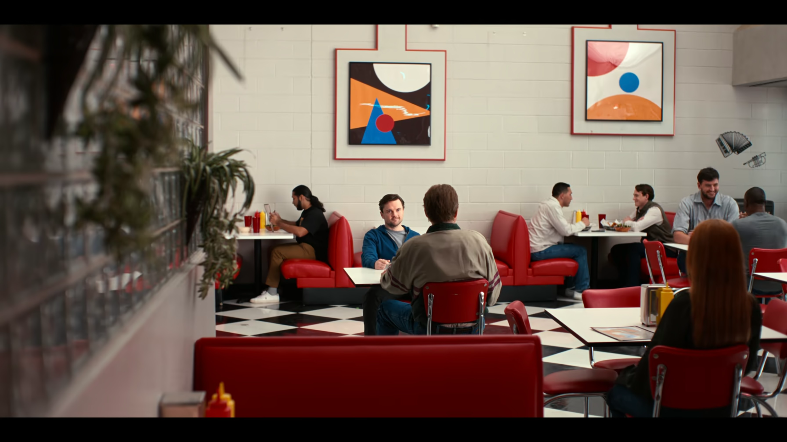 Interior of a retro-style diner with red booths and chairs, customers dining and talking, abstract artwork on white brick walls, black and white checkered floor, and a few potted plants.