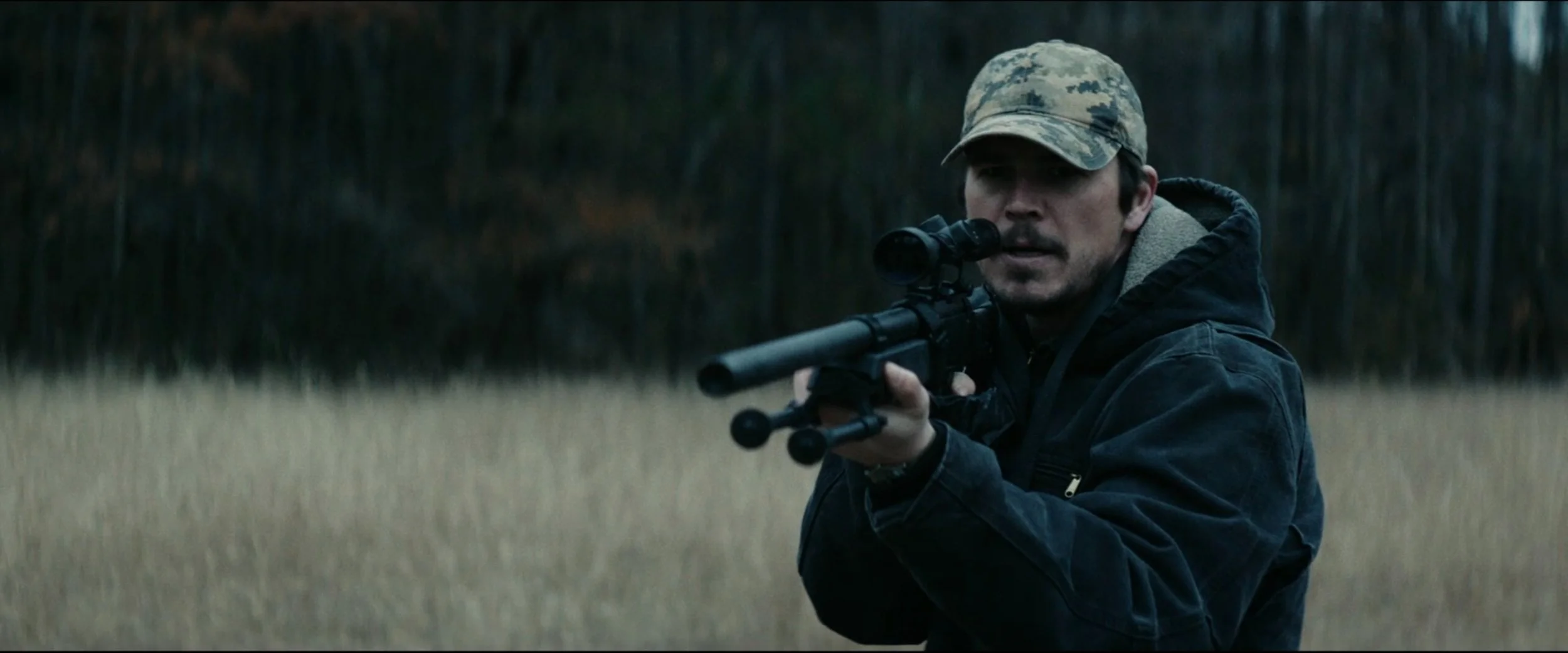 A man in a camouflage cap aimed with a rifle in a field with trees in the background.