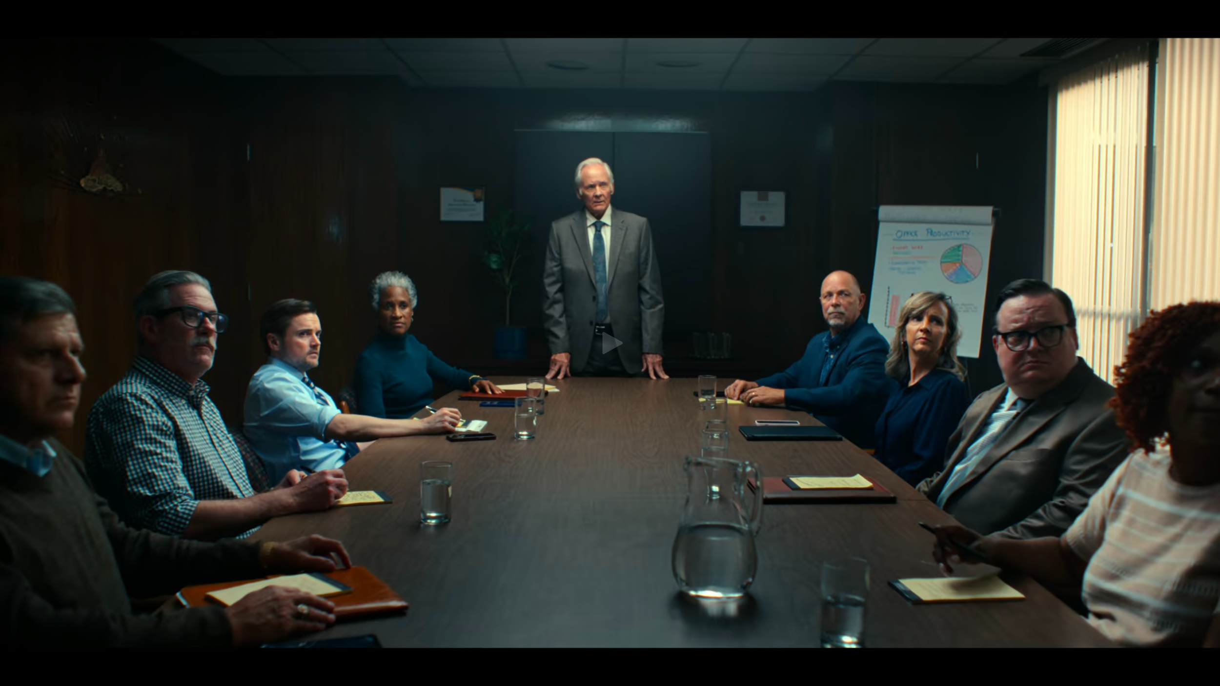 A group of nine diverse people in a boardroom, seated around a large table, with one man standing at the head of the table. The room has wooden walls, a window with blinds, and a whiteboard with charts and notes.