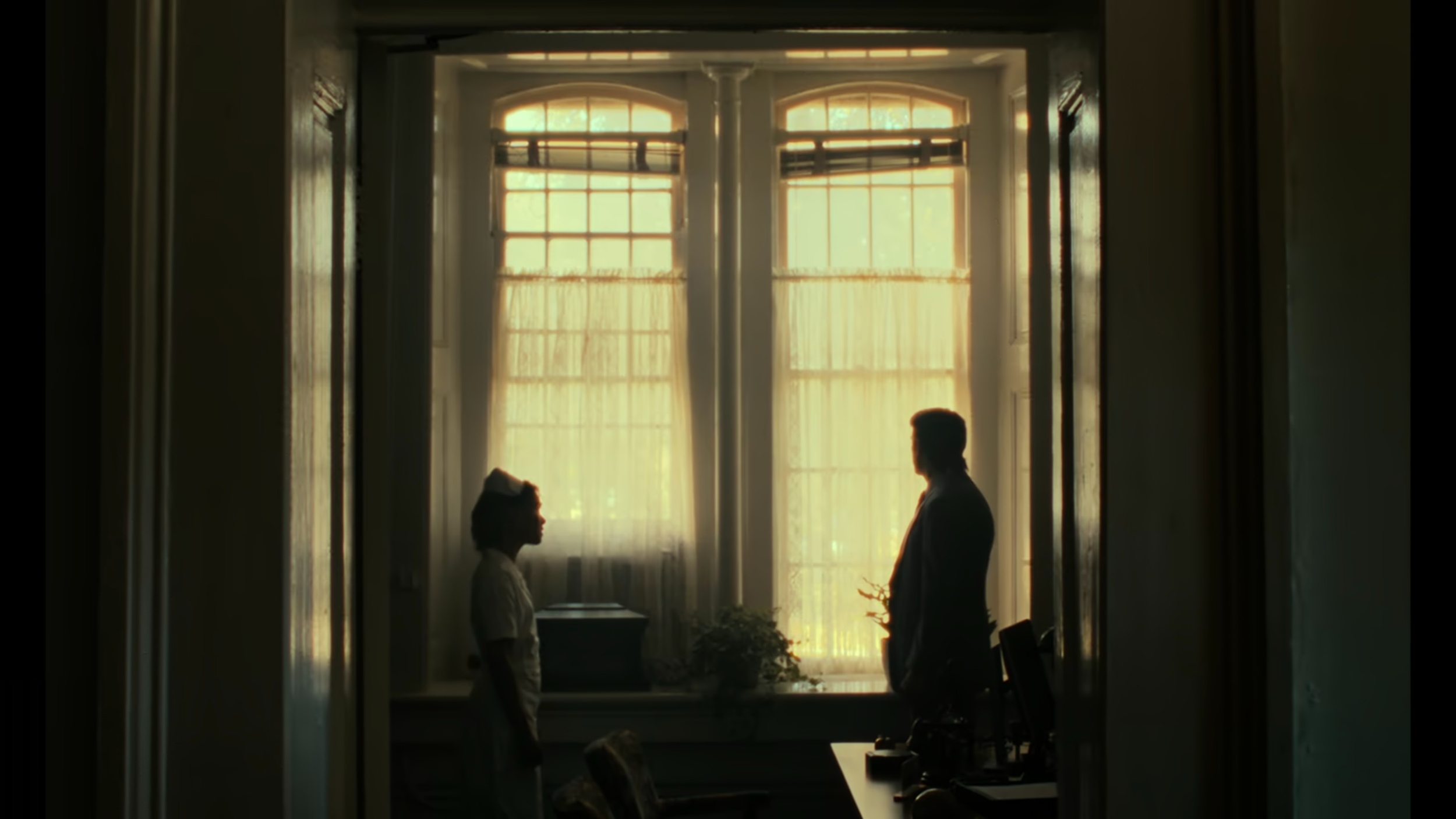 A silhouette of a nurse and a man standing and facing each other in a room with large windows and curtains, backlit by sunlight.