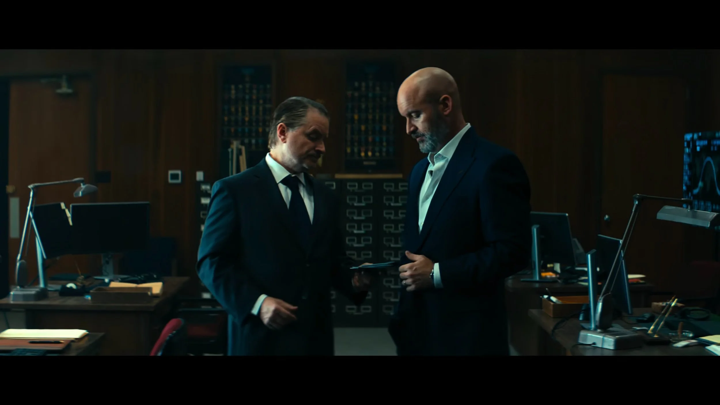 Two men in suits standing in an office, looking at a document.