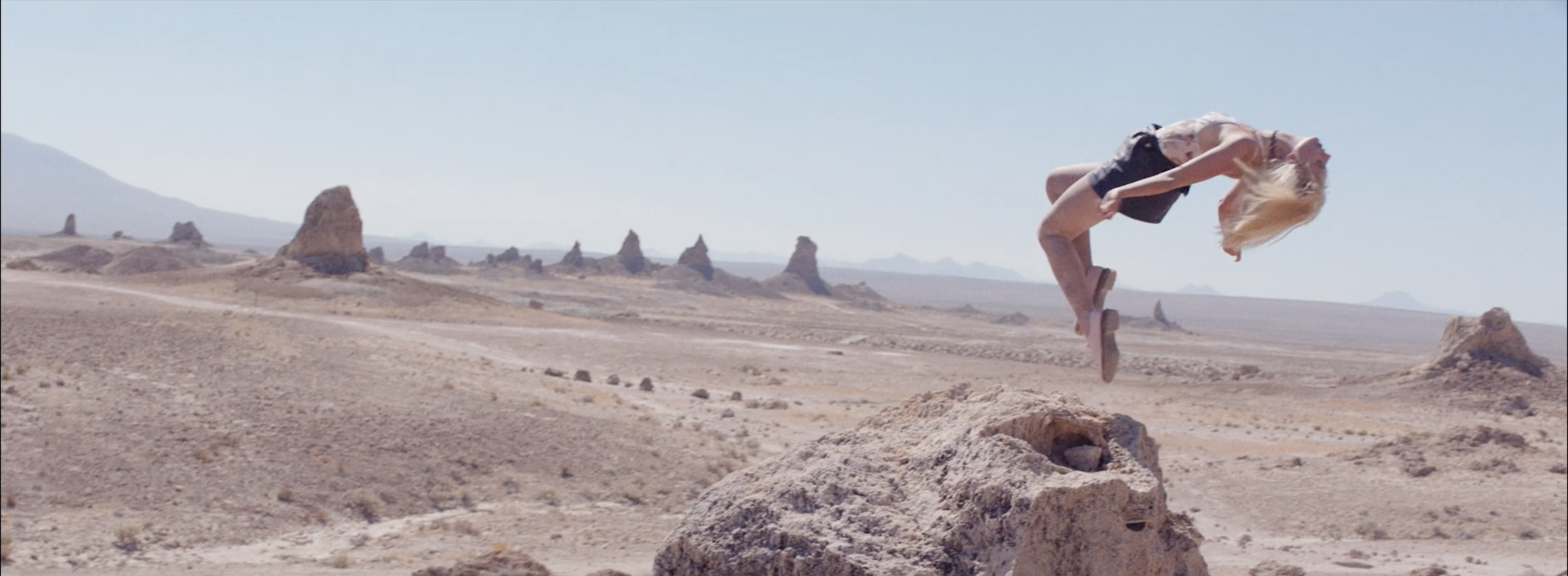 A person with blonde hair jumping high in a desert landscape with rocky formations and distant mountains under a clear blue sky.