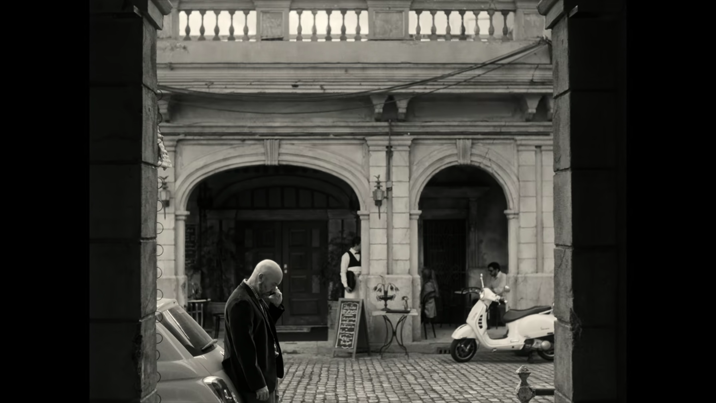 A black and white photograph of a city street scene through a stone archway. A man with a bald head is standing near a white car, talking on a cell phone. In the background, a woman is standing outside a building, and another woman is riding a scooter. There is a small table with a menu board and a statue outside the building.
