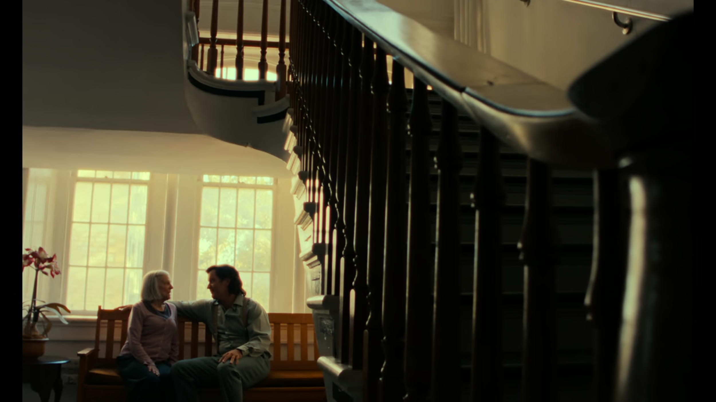 Two people sitting on a wooden bench in front of a large window with divided panes, with the staircase railing partially blocking the view.